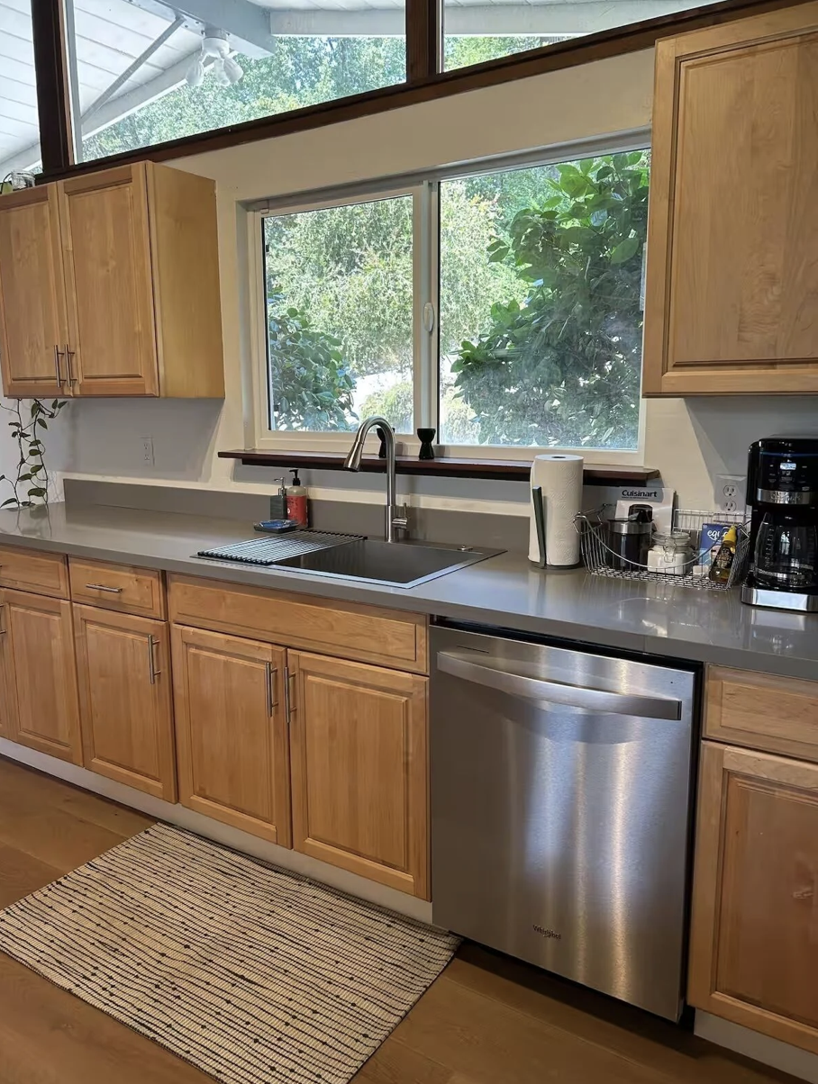 Kitchen with wooden cabinets, a window over the sink with greenery outside, and various small appliances on the countertop including a coffee maker, paper towels, and a dish drying rack.