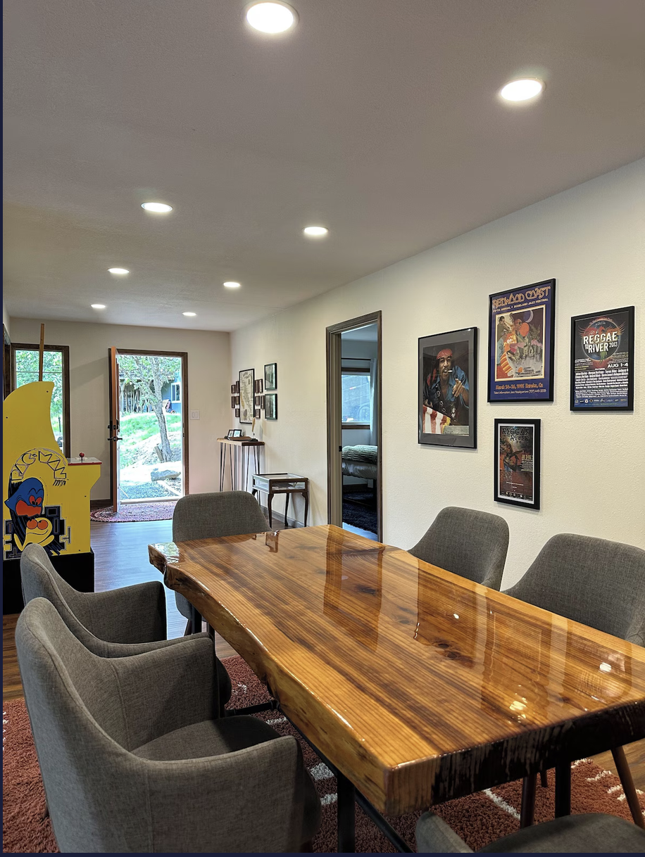 A dining room with a polished wooden table surrounded by six gray upholstered chairs, a doorway leading to a bedroom, wall art featuring posters, a small side table, and an open door with a view of the outdoors.
