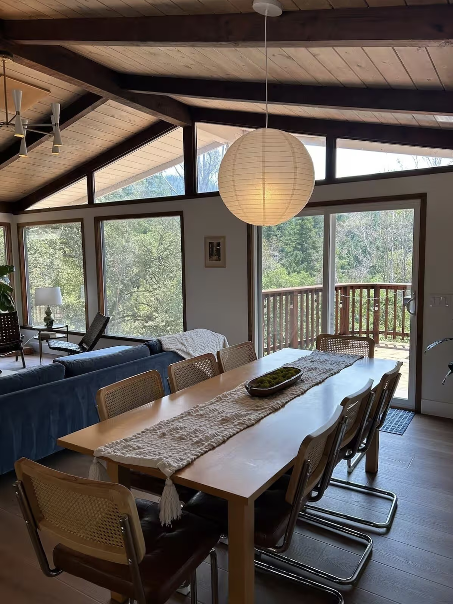 This is a dining and living room area with large windows showing trees outside, a wooden ceiling with beams, a round paper lantern light fixture, a long wooden dining table with a cloth runner and eight chairs, a blue sofa with a throw blanket, a sid