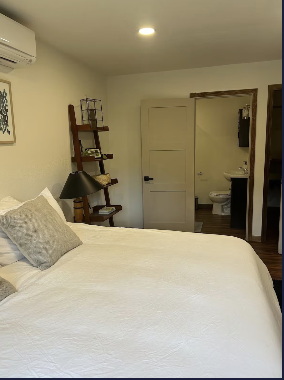 A cozy bedroom with a large white bed, gray pillows, a black bedside lamp, a wooden ladder shelf, and an en-suite bathroom visible through an open door.