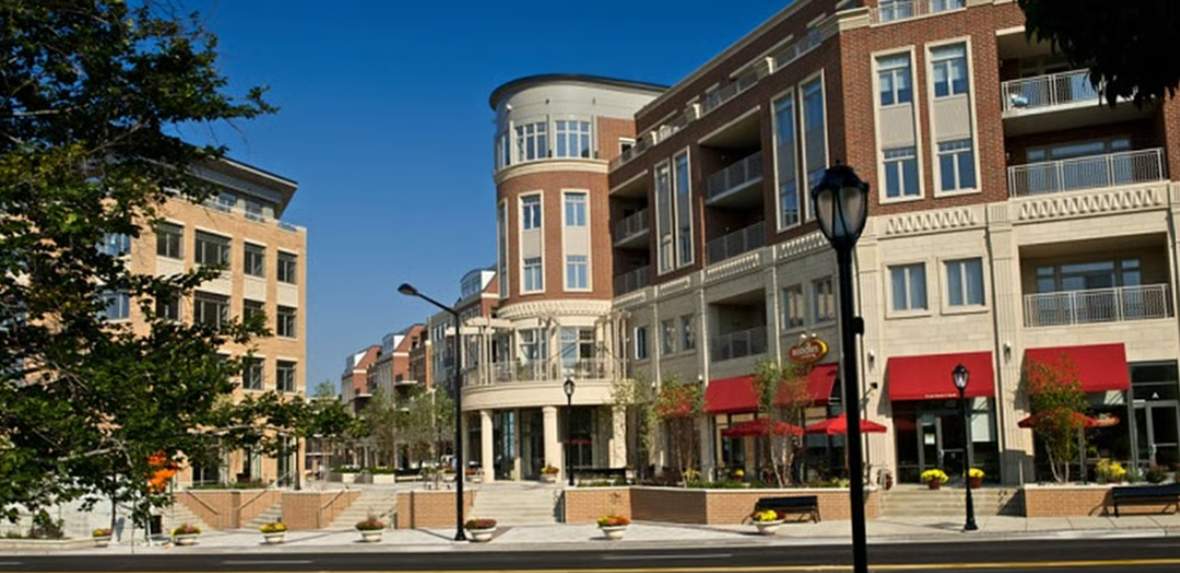 A city street lined with modern, multi-story buildings with balconies and red awnings, trees, benches, lampposts, and potted plants, under a clear blue sky.
