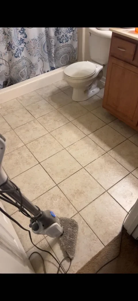 A bathroom with a toilet, wooden vanity, tiled floor, and a vacuum cleaner in the foreground.
