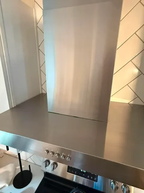 Stainless steel stovetop with a vent above it, in a modern kitchen with tiled walls.