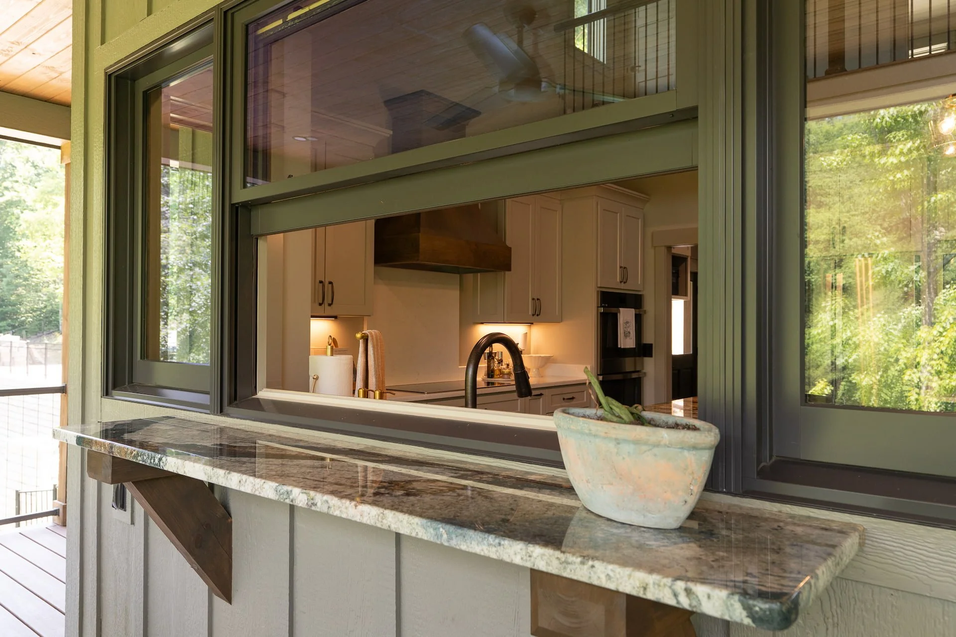 View of a kitchen through an open window, showing white cabinets, a black faucet, and outdoor greenery.