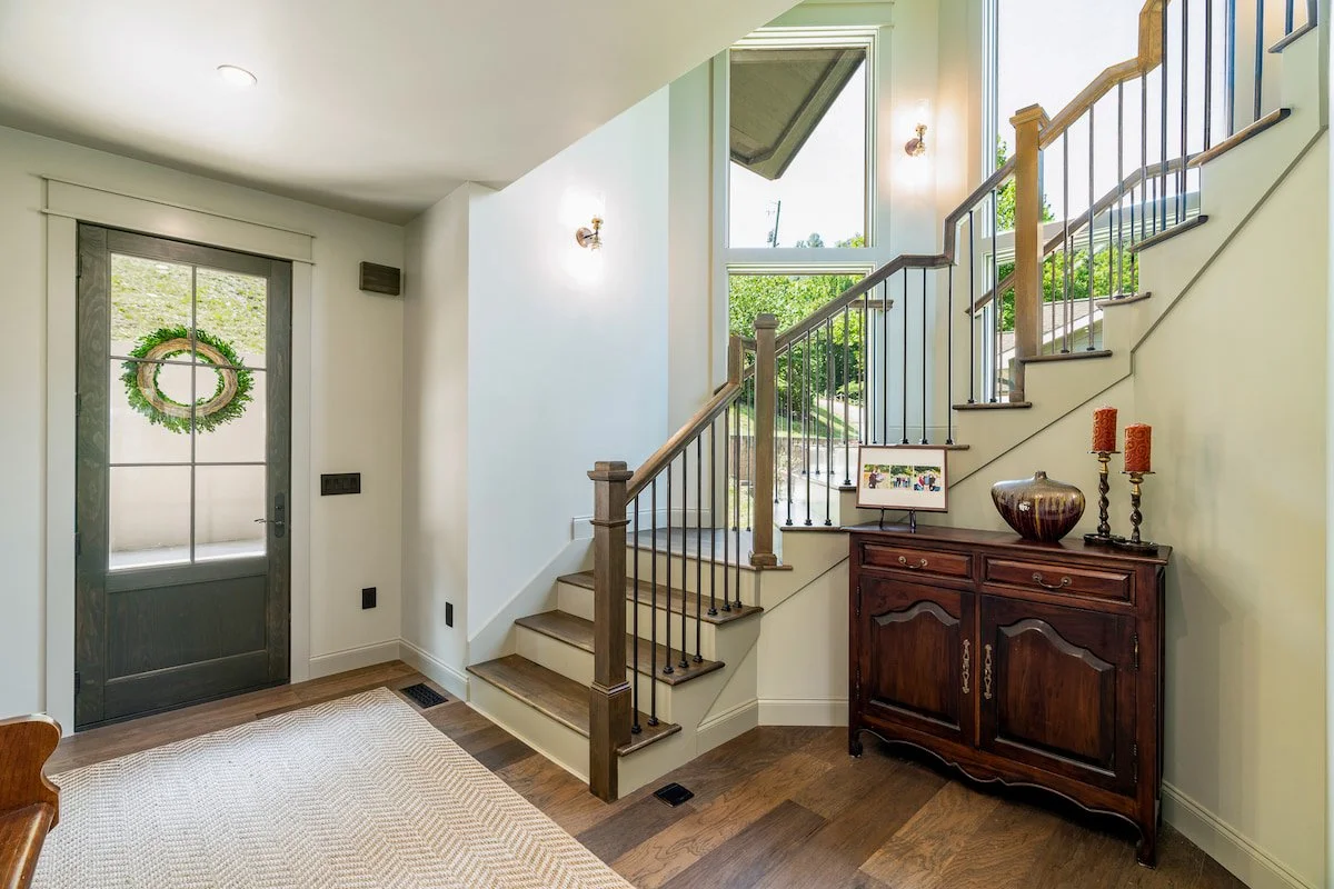 Interior view of a home's front entryway featuring a staircase, a wooden sideboard with decorative items, and a window with outdoor view.