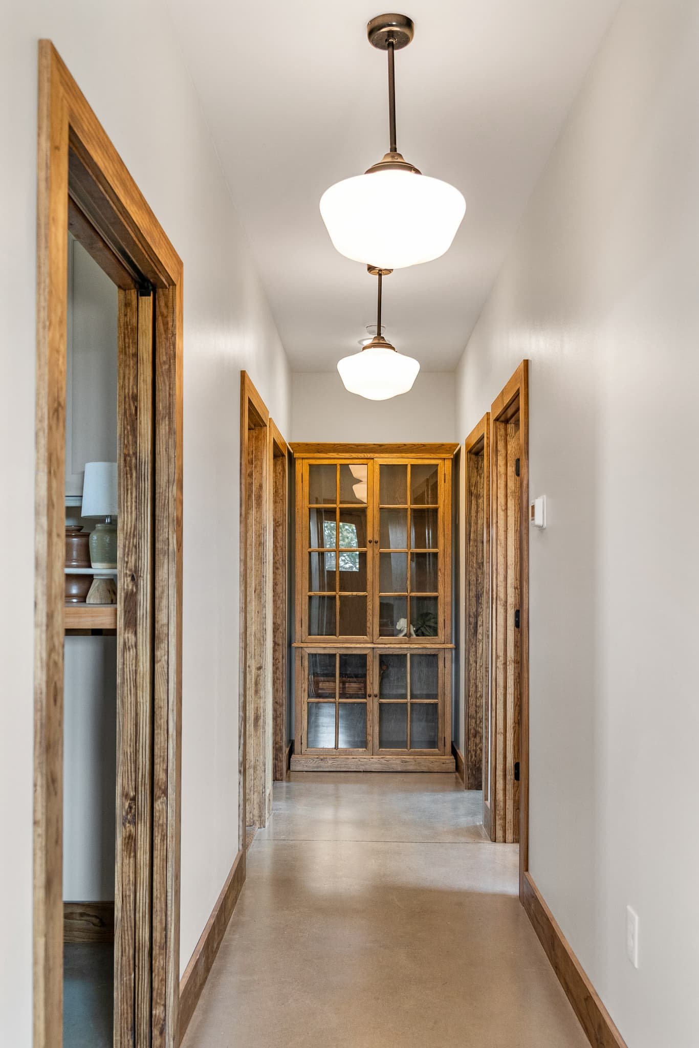 A narrow hallway with white walls and wooden trim, featuring two hanging ceiling lights and glass-paned wooden double doors at the end.