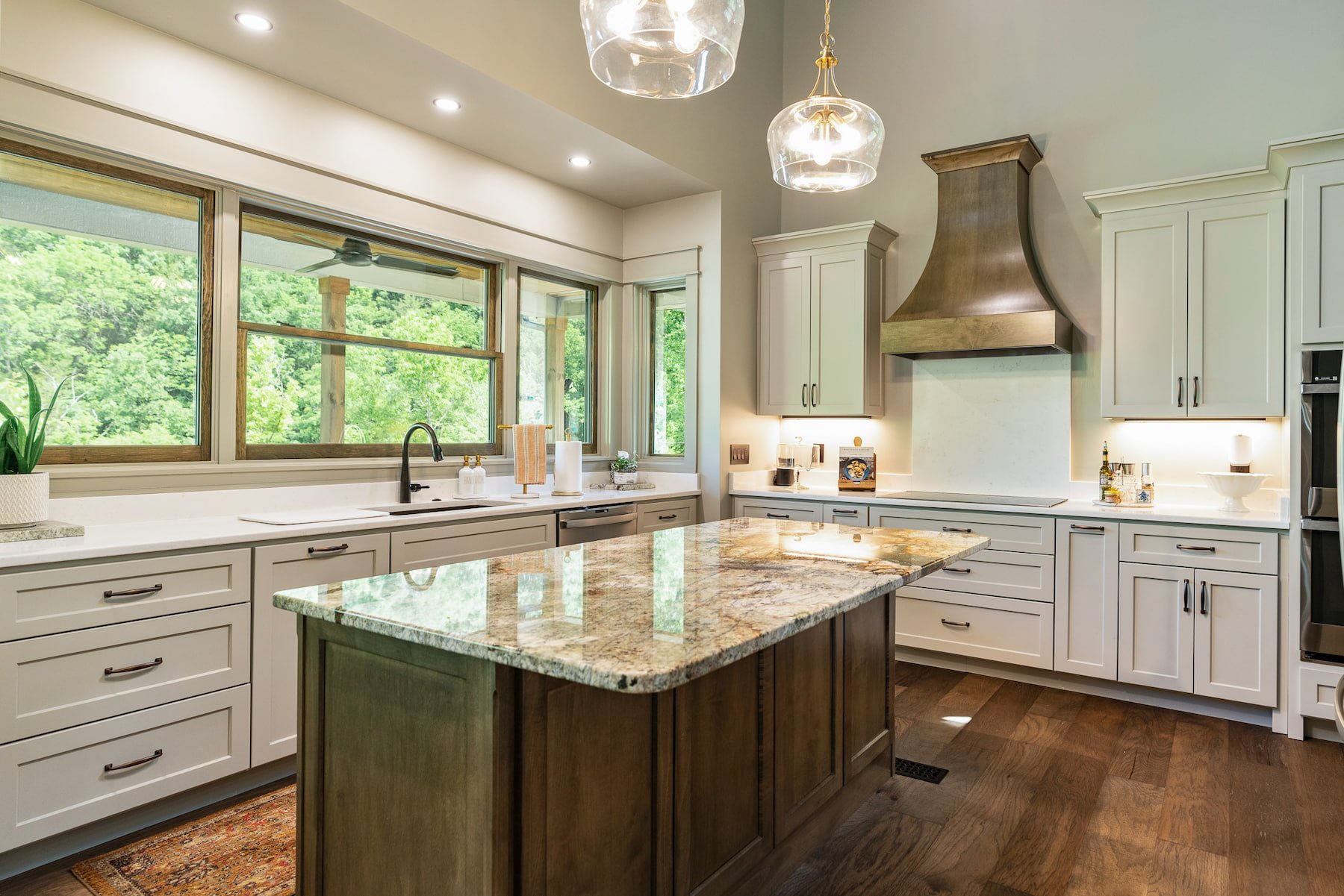Modern kitchen with white cabinets, a marble countertop island, large windows overlooking greenery, a stainless steel range hood, pendant lights, and hardwood floors.
