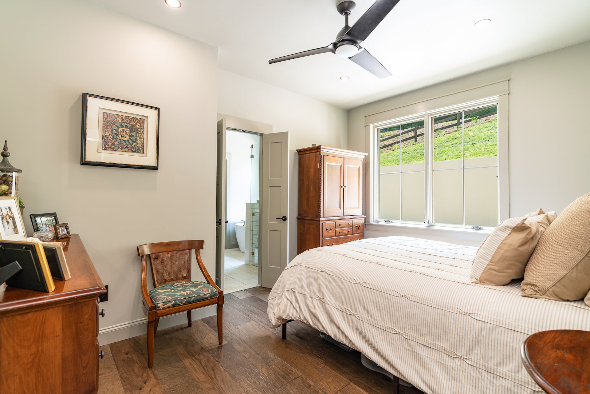A bedroom with a bed, window, wooden armoire, ceiling fan, and a small bathroom visible through an open door.