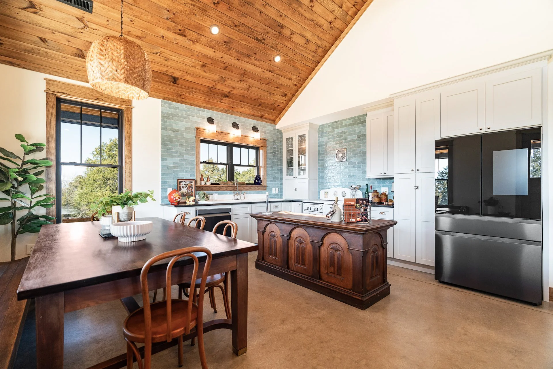 A modern kitchen with a wooden ceiling, white cabinets, a black refrigerator, and blue tiled backsplash. There is a wooden island, a dining table with chairs, and various decorative items on the countertops and table. Large windows let in natural light and showcase greenery outside.