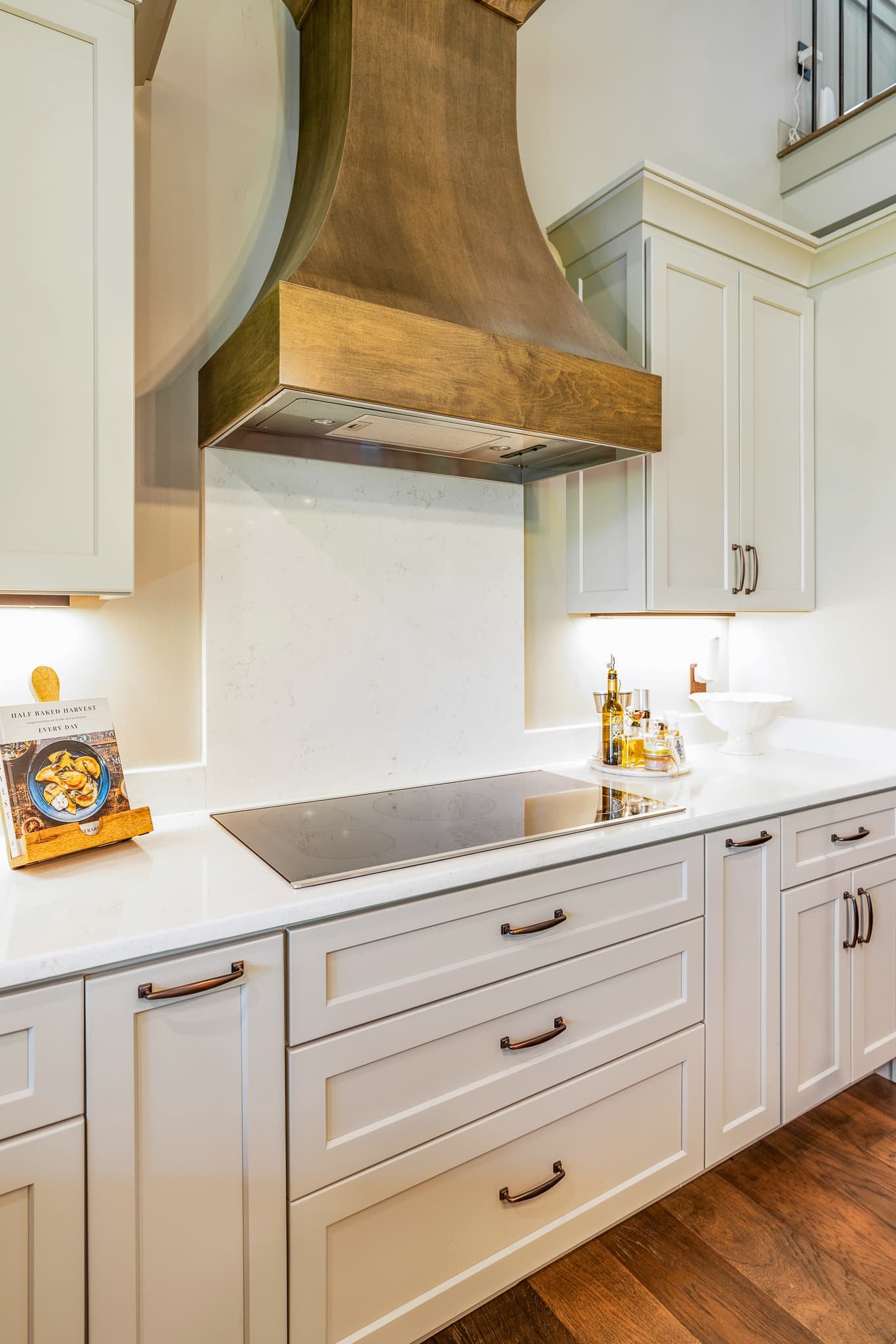 White kitchen cabinets with bronze handles, a black electric stovetop, a white countertop, a small cookbook or magazine on a stand, and a collection of olive oil bottles and a white bowl on the countertop. There are wood floors.