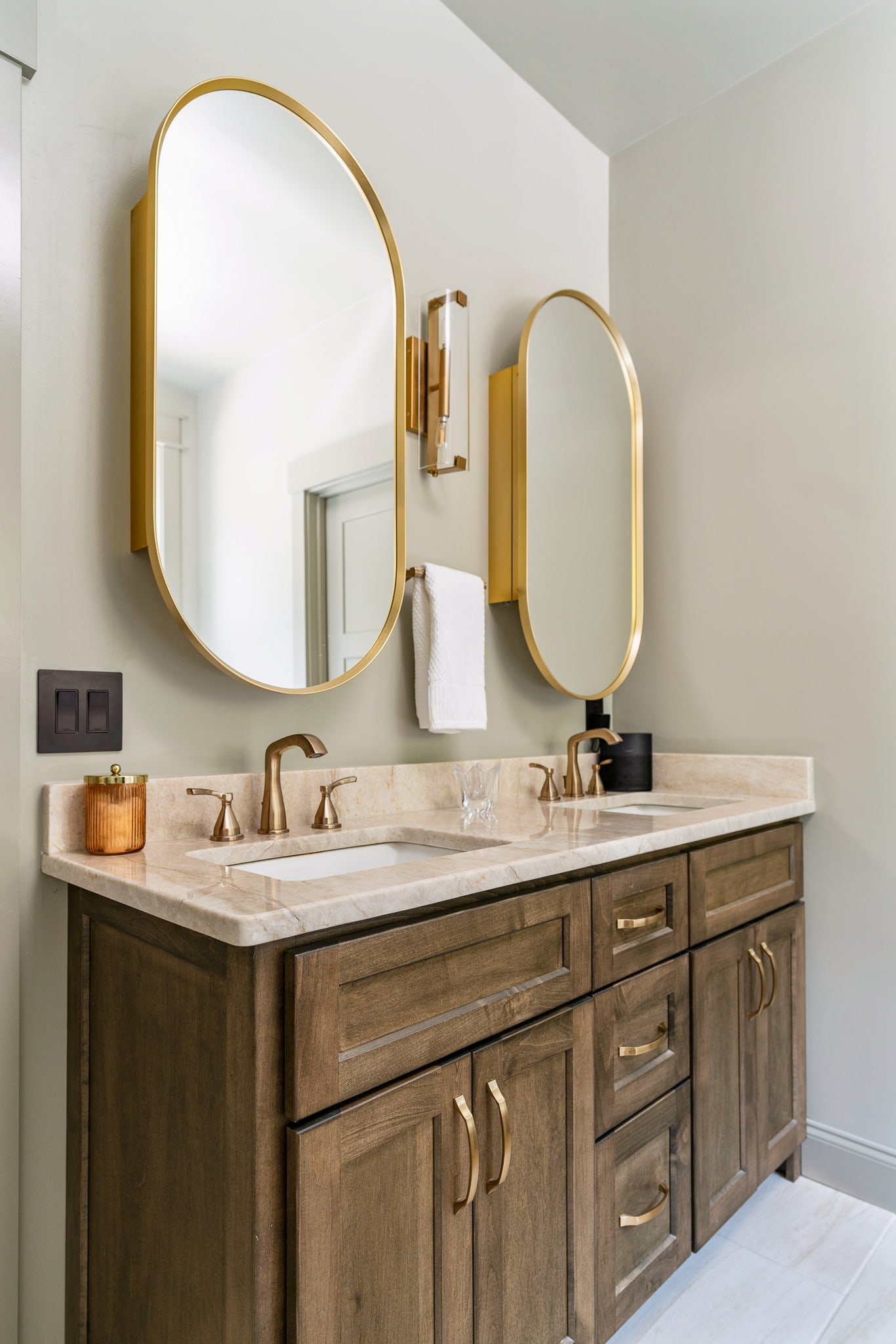 Bathroom vanity with dark wood cabinet, beige marble countertop, gold hardware, two oval mirrors with gold frames, and a wall-mounted light fixture.