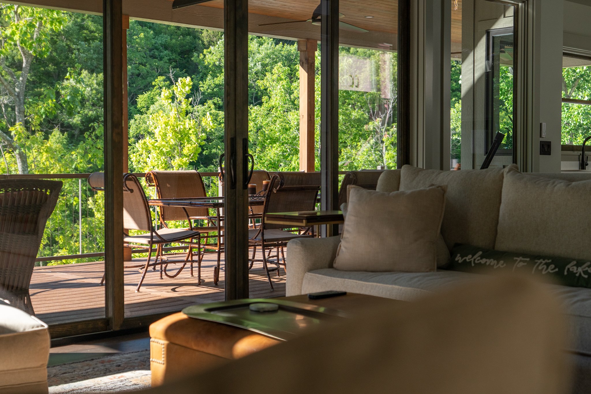 Living room with a sofa, a throw pillow, and a coffee table, looking out through sliding glass doors to a balcony with chairs and lush green trees in the background.