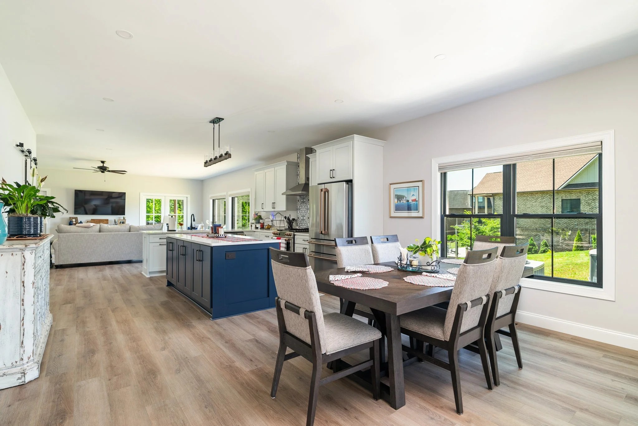 Open-concept kitchen and dining area with large windows, white cabinets, a blue kitchen island, and a wooden table with chairs.