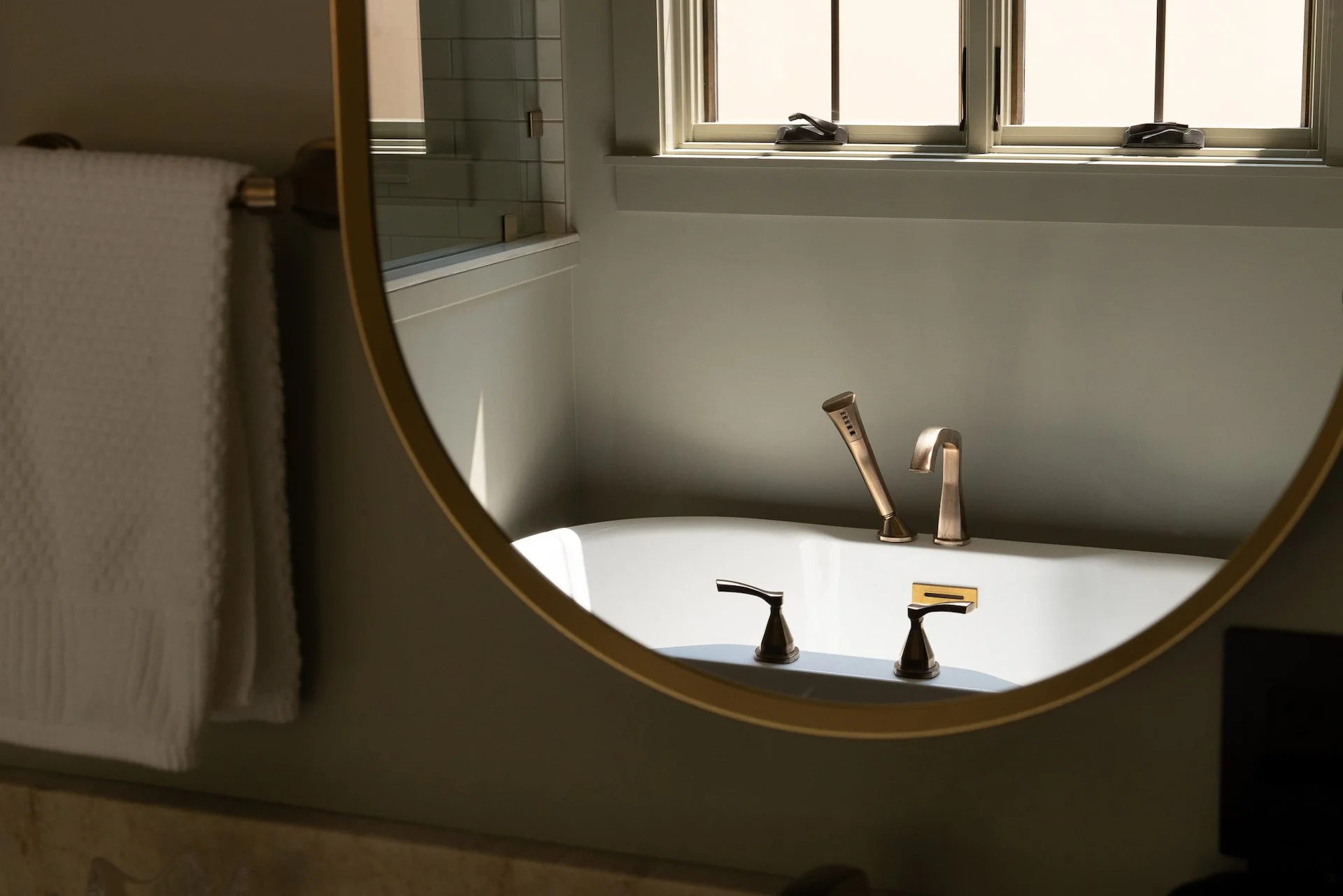 Bathroom sink with three modern faucets reflected in a circular mirror, overlooking a window with partially opened blinds.