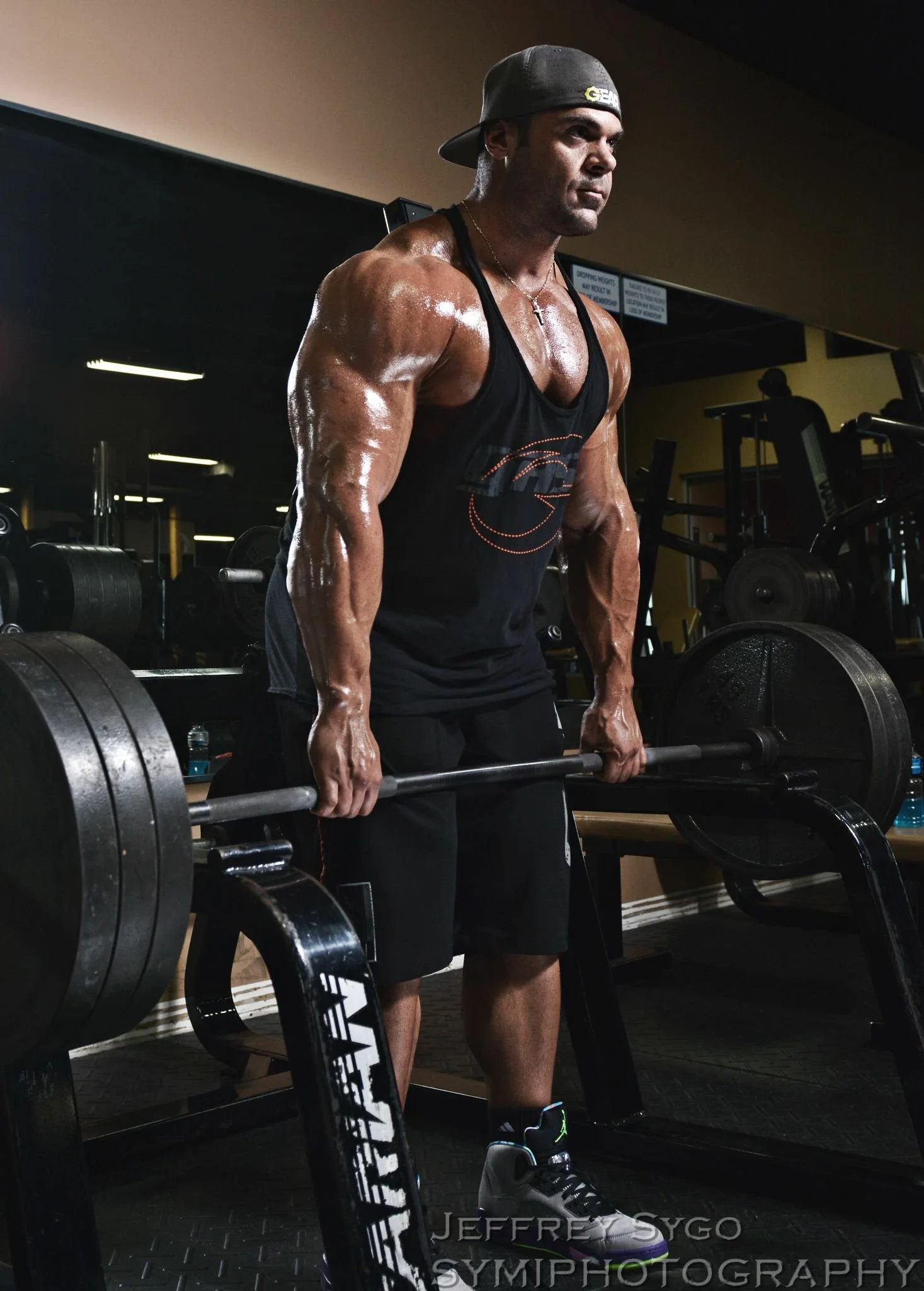 A muscular man lifting a heavy barbell in a gym, wearing a black tank top, black shorts, a backwards cap, and sporting a necklace with a cross, with gym equipment and weights in the background.