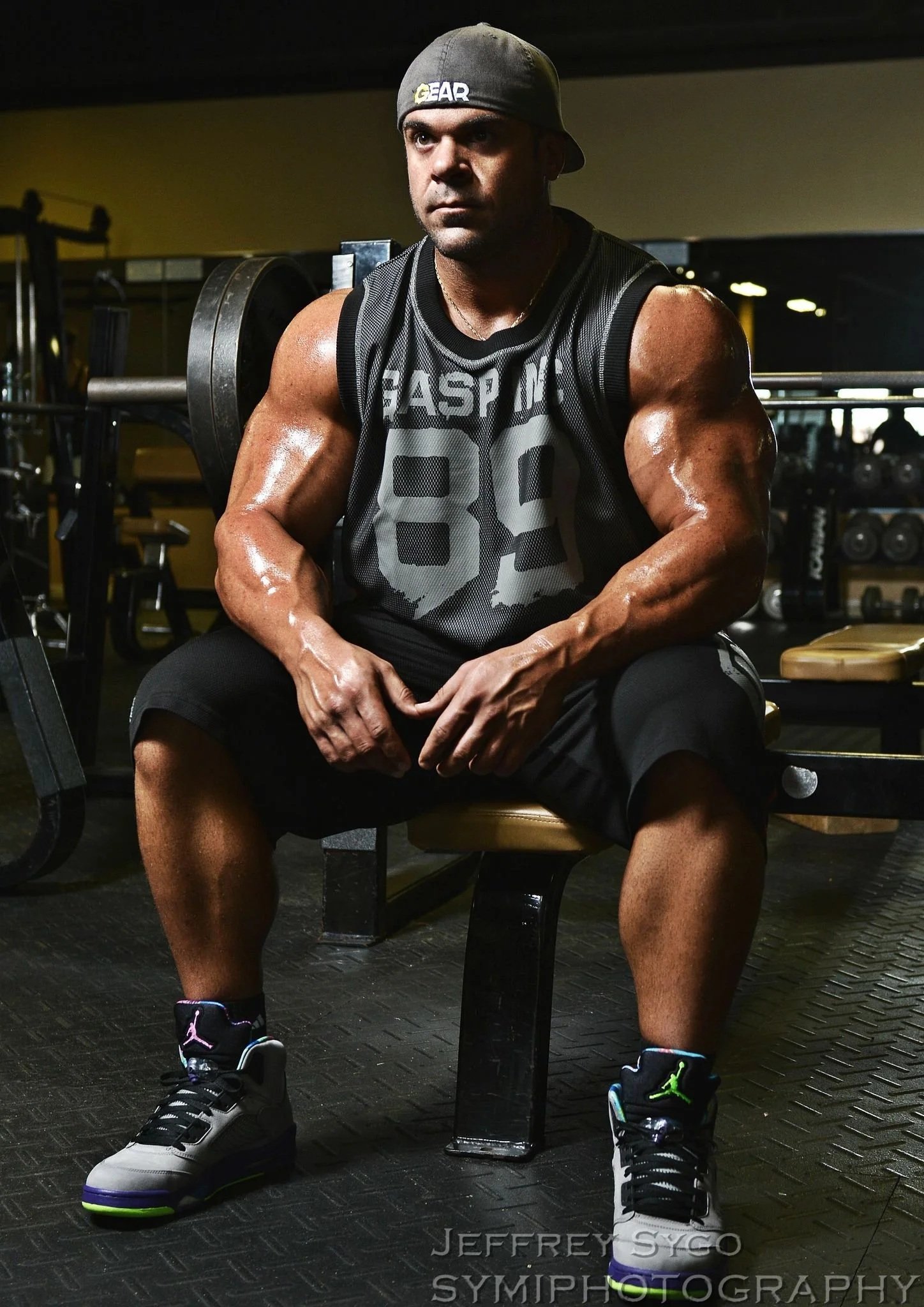 A muscular man wearing a gray cap backwards, a black athletic sleeveless shirt, black shorts, and black, white, purple, and green sneakers sits in a gym with weightlifting equipment in the background.