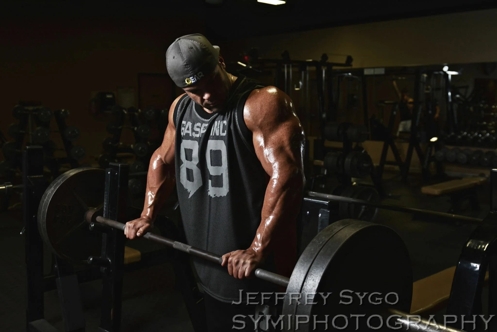 A muscular man wearing a gray cap and a black sleeveless shirt with the number 89, lifting a barbell in a gym with multiple dumbbells and weights in the background.