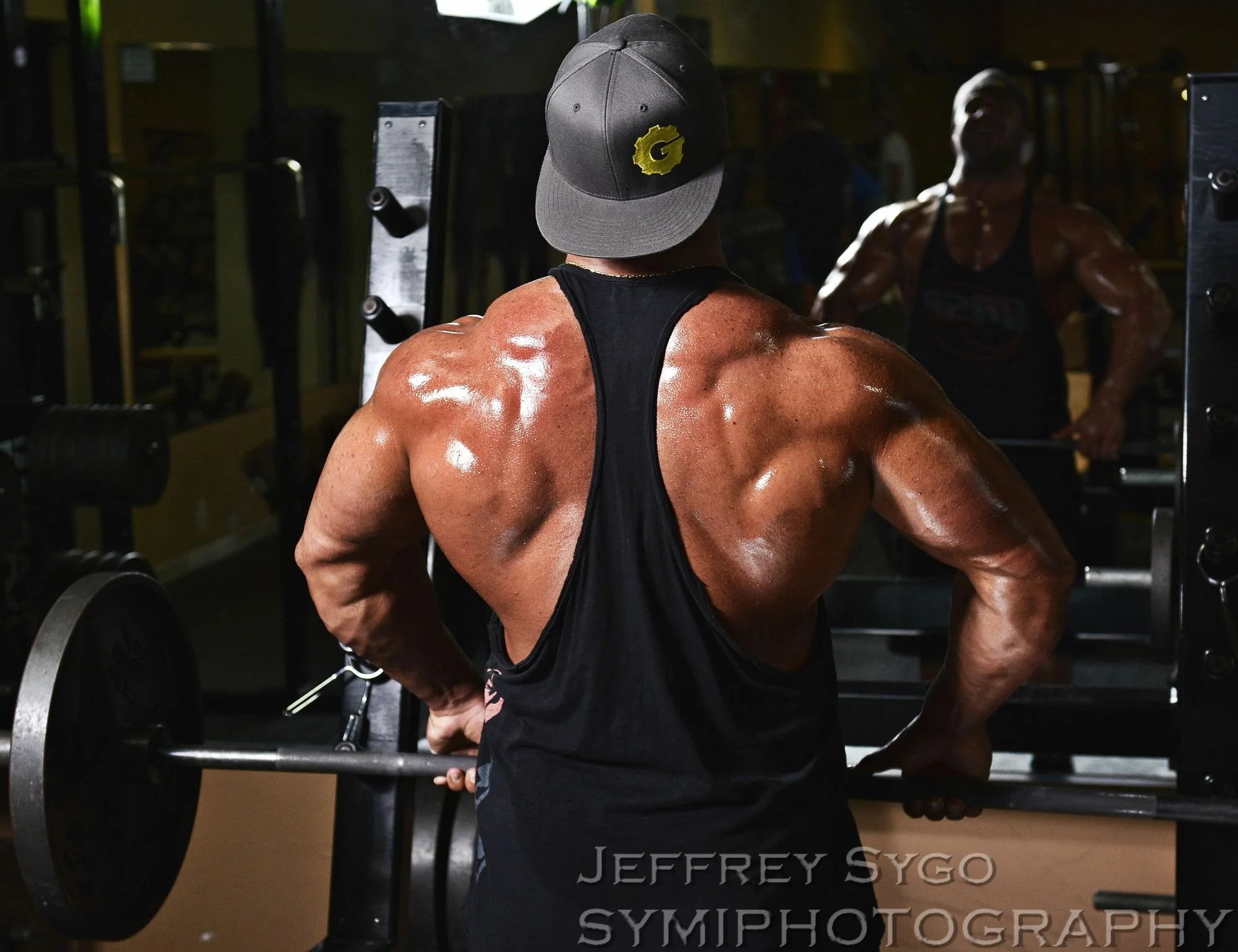A muscular man in a black tank top and gray cap with a yellow logo, standing in front of a mirror in a gym, holding a barbell, with a reflection of his upper body and face visible in the mirror.