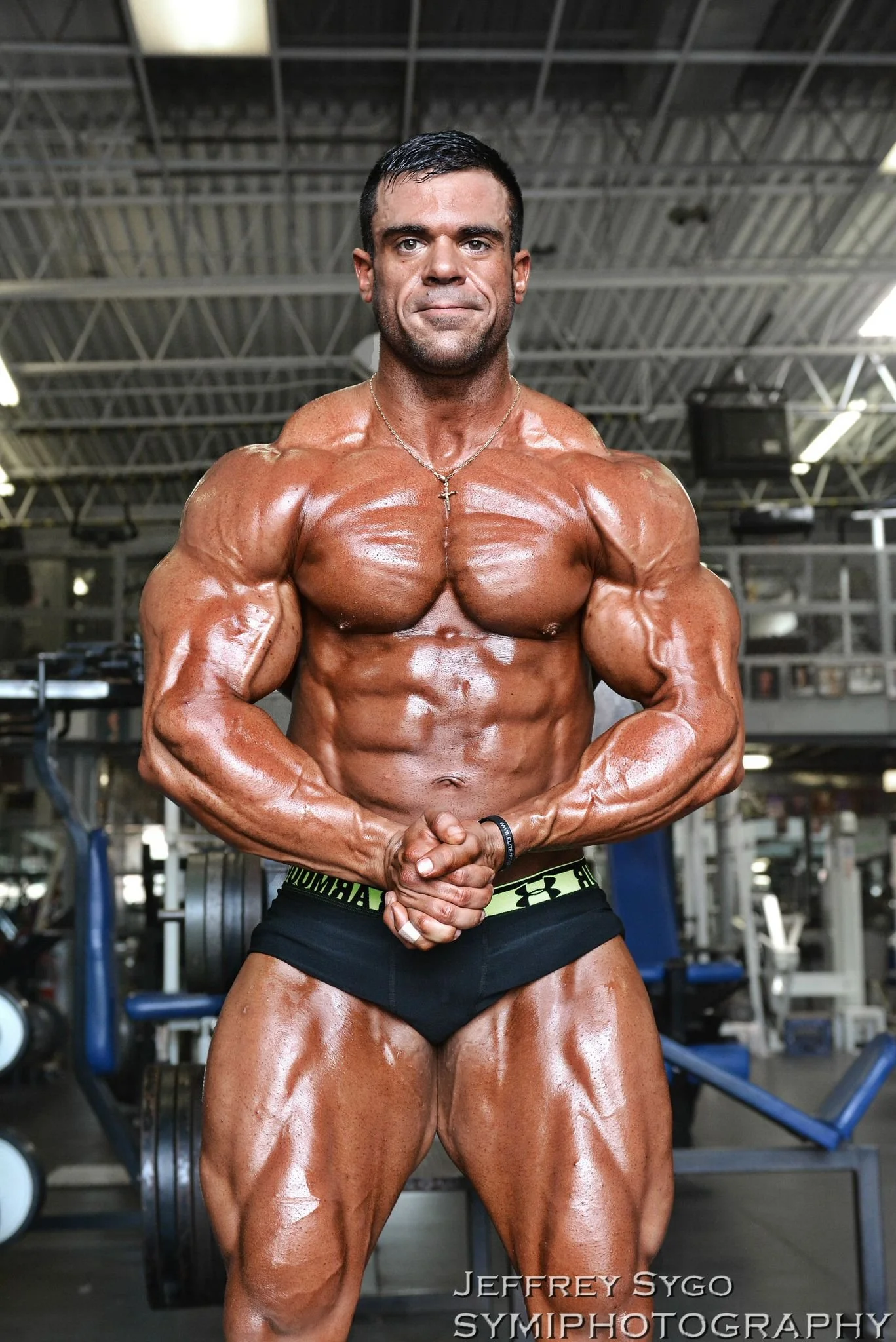 A muscular man with short dark hair and a cross necklace posing in a gym, flexing his arm muscles. The gym has a high ceiling with grid patterns and exercise equipment in the background.