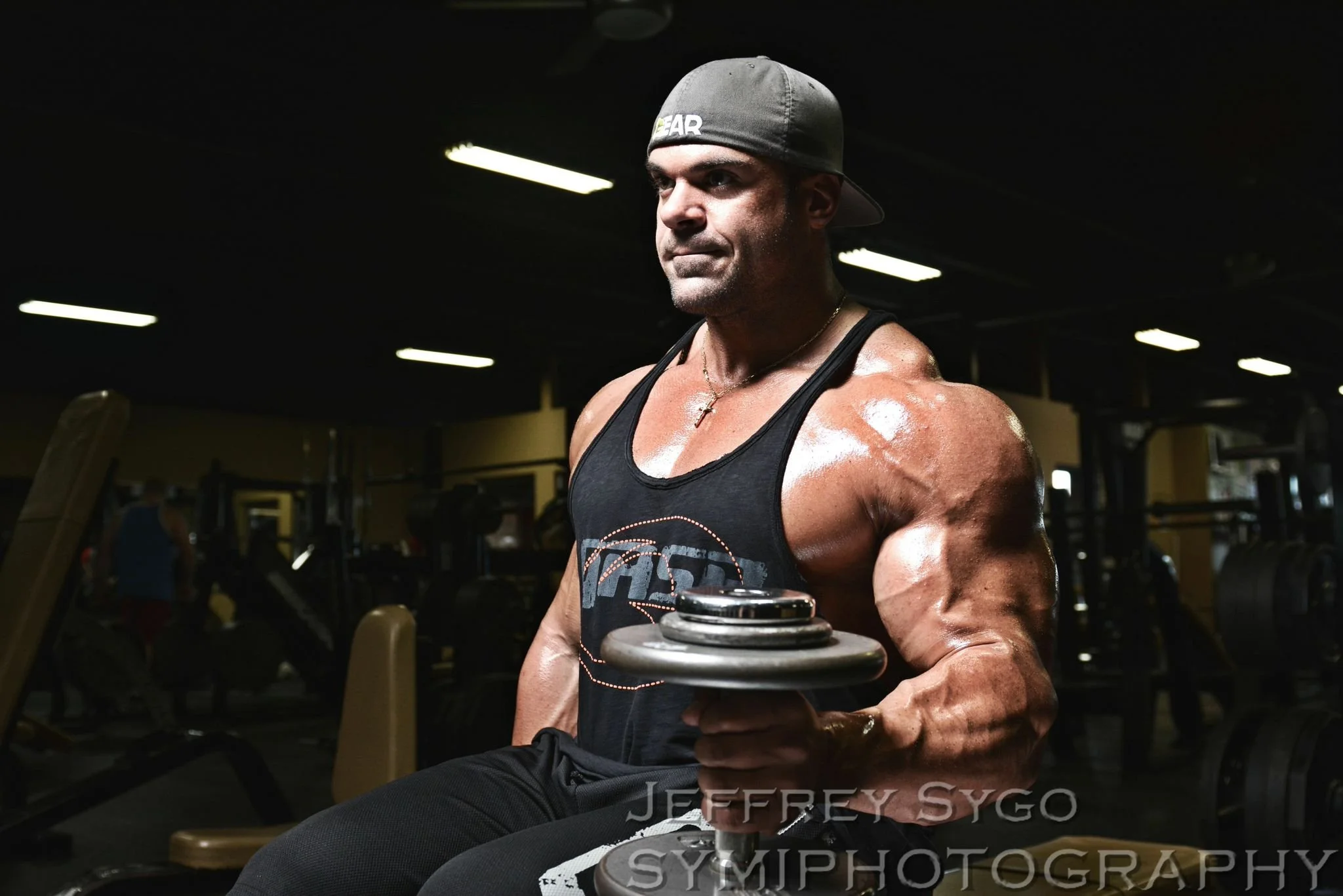 A muscular man wearing a gray cap backwards and a black tank top, holding a dumbbell in a gym with dim lighting and exercise equipment in the background.