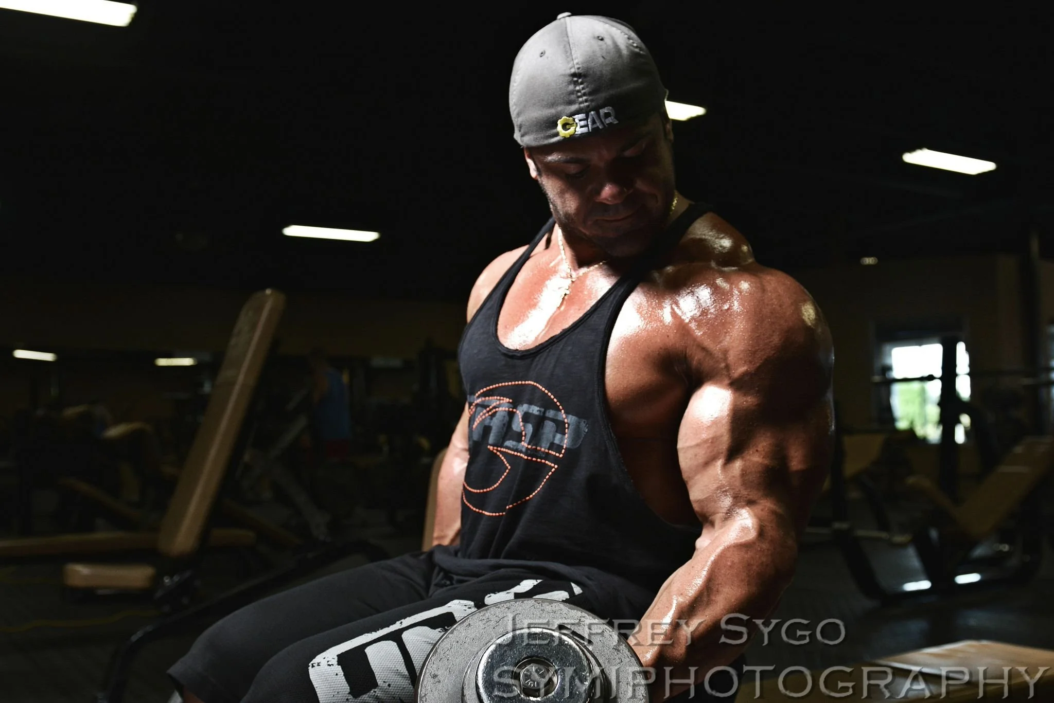 A muscular man wearing a gray cap and black tank top is lifting a dumbbell in a gym, with sweat on his face and shoulders, illuminated by overhead lights.