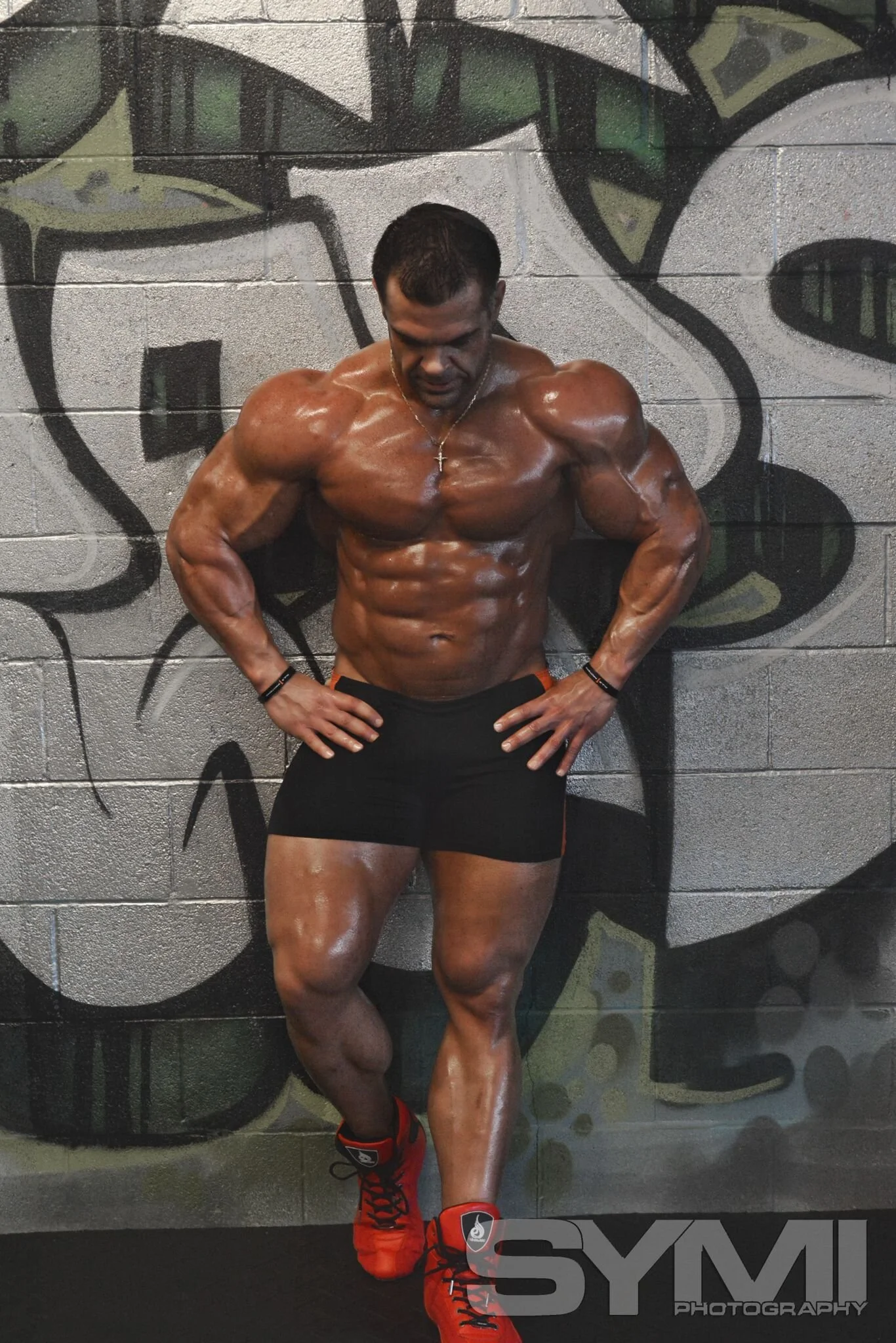 A muscular man standing against a graffiti-covered wall, wearing black shorts, red and black athletic shoes, and a silver cross necklace, with his hands on his hips and head tilted downward.