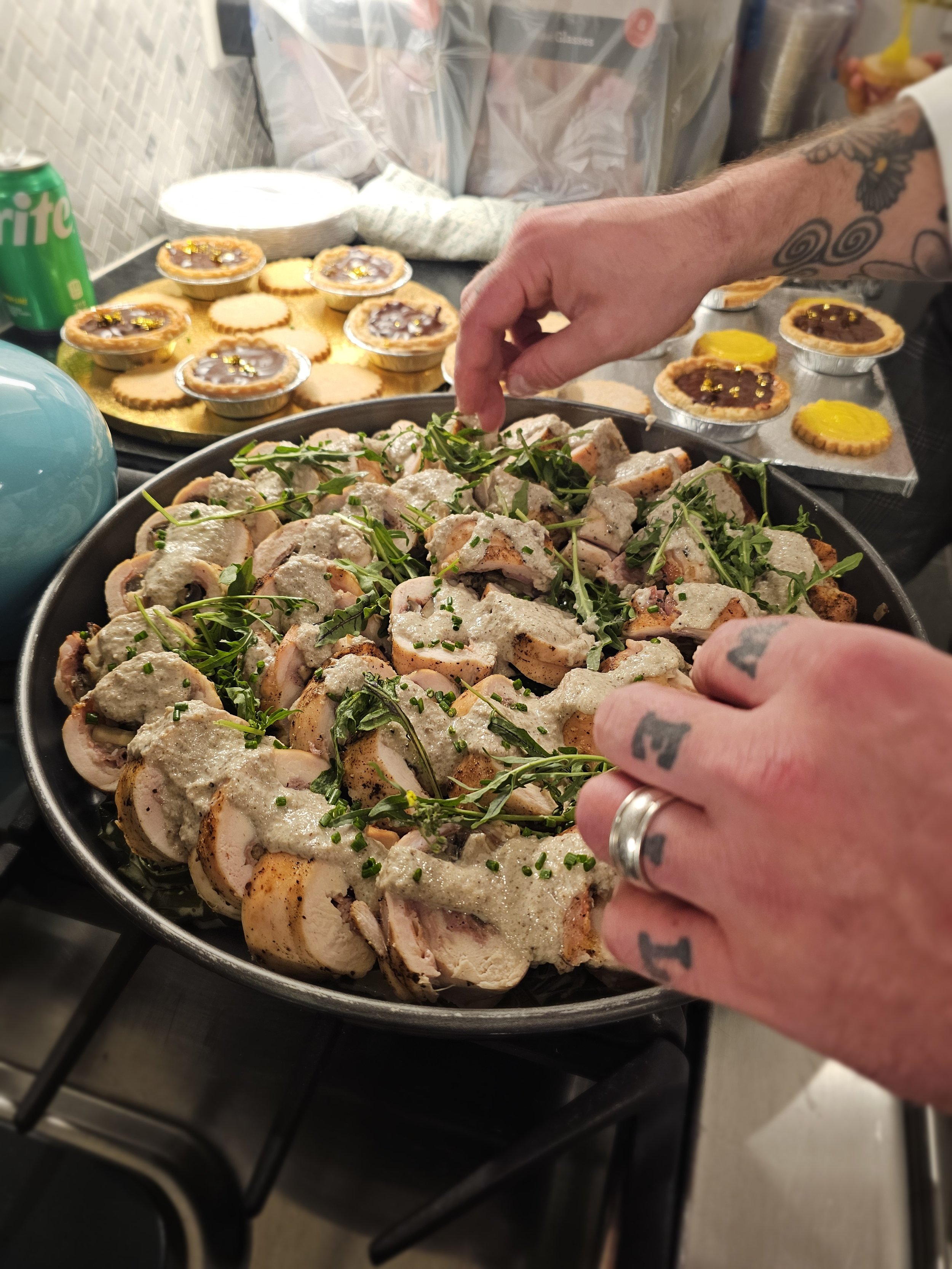 A person with tattoos on their hand is garnishing a platter of sliced cooked chicken with chopped herbs, with a background of holiday desserts including mini pies and cookies.