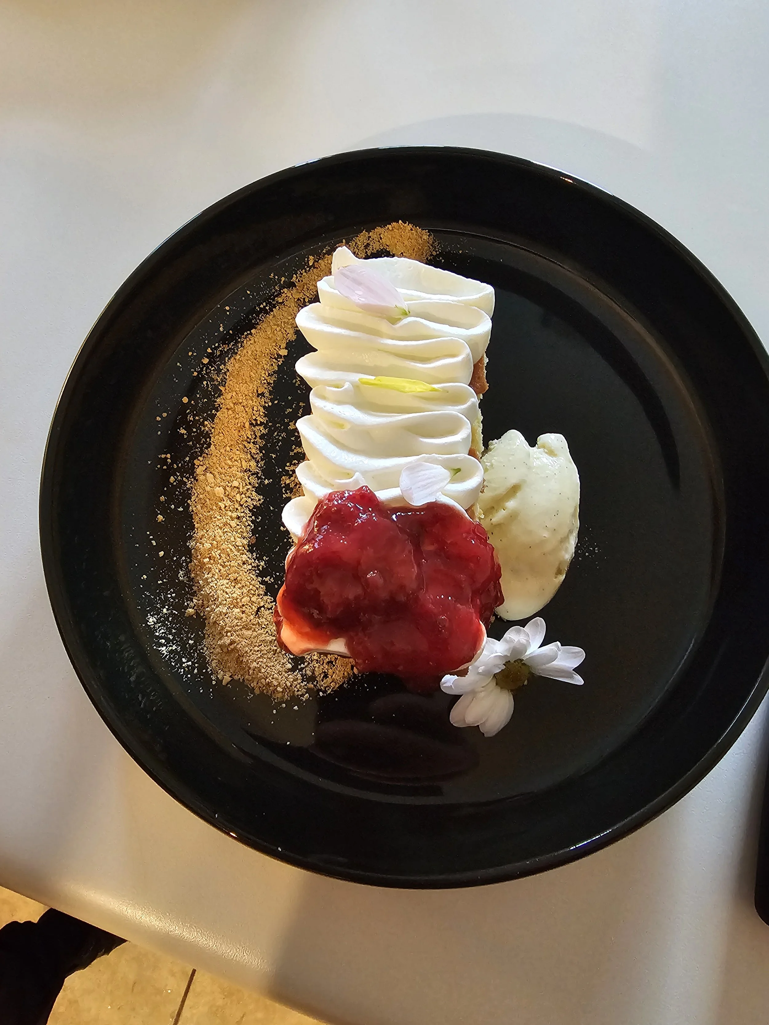 A dessert plate with whipped cream, strawberry sauce, a flower, and a graham cracker crust decorated with a flower petal and sprinkled with crumbled cookie or biscuit.