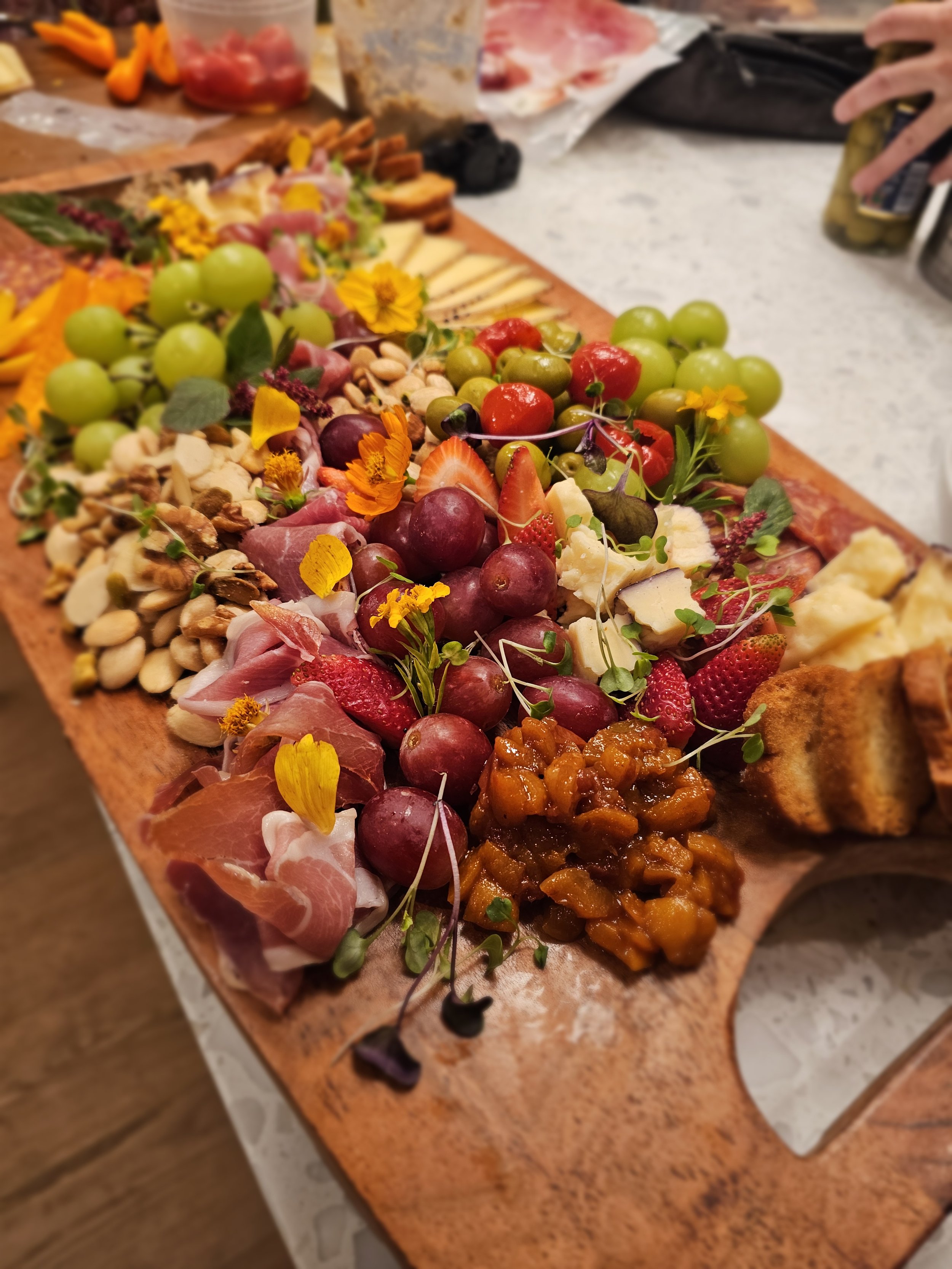 A wooden charcuterie board with a variety of cheeses, fresh grapes, strawberries, sliced meats, nuts, and garnished with edible flowers, set on a table.