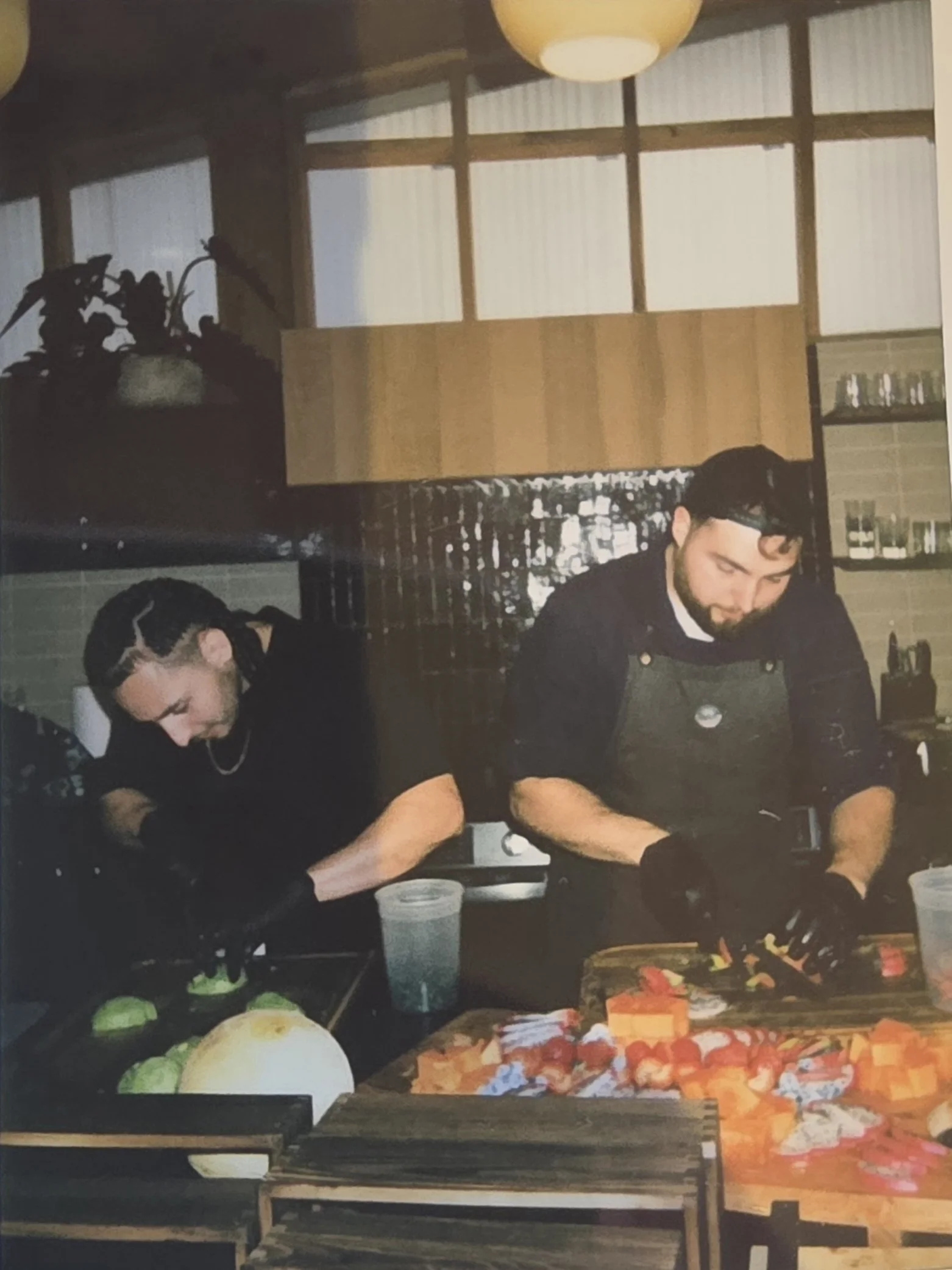 Two men preparing food in a rustic kitchen with wooden shelves and large windows. The counter displays various vegetables and ingredients.