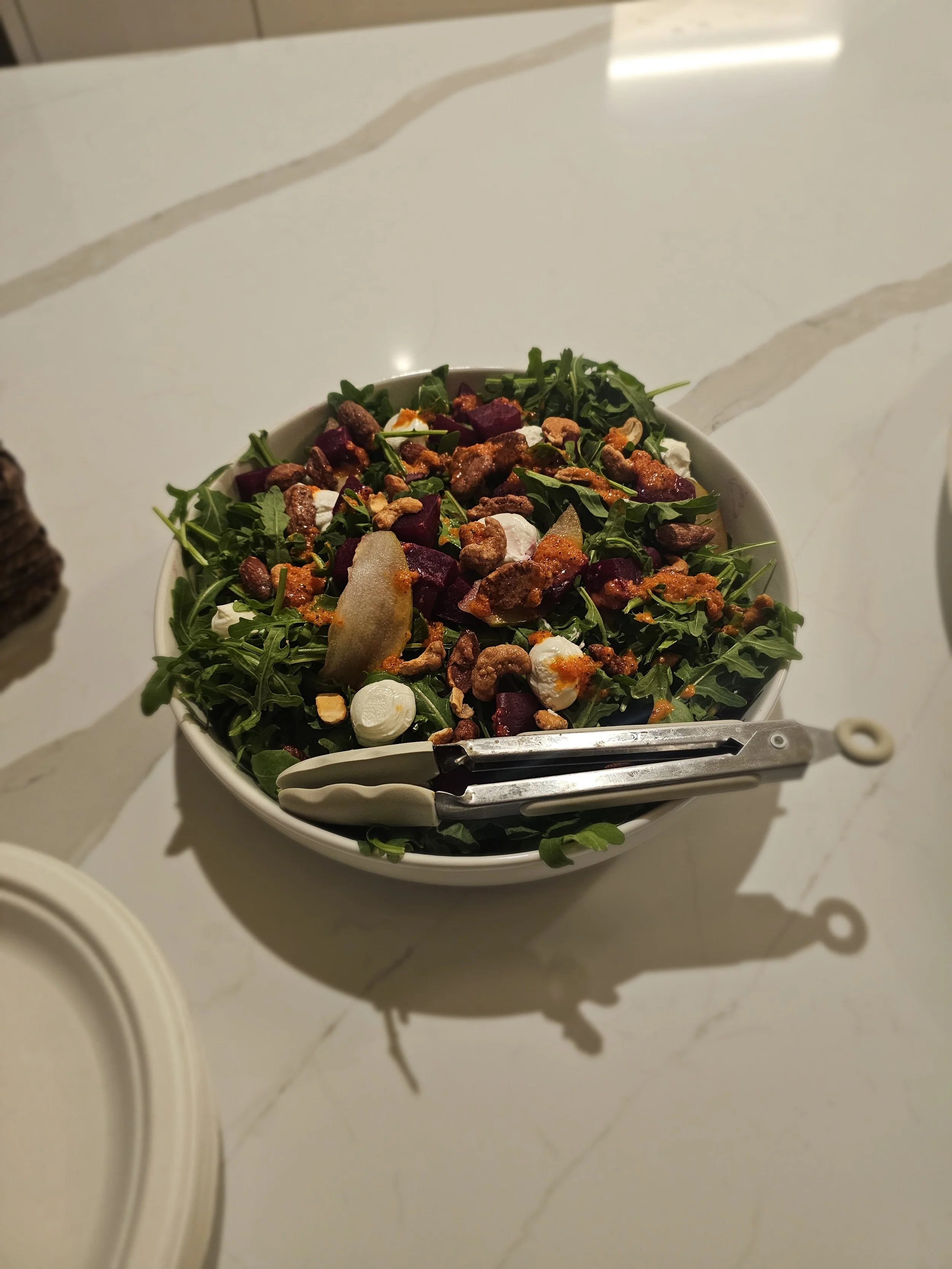 A white bowl filled with a fresh salad containing arugula, roasted beets, goat cheese, walnuts, and a dressing, placed on a white marble countertop.
