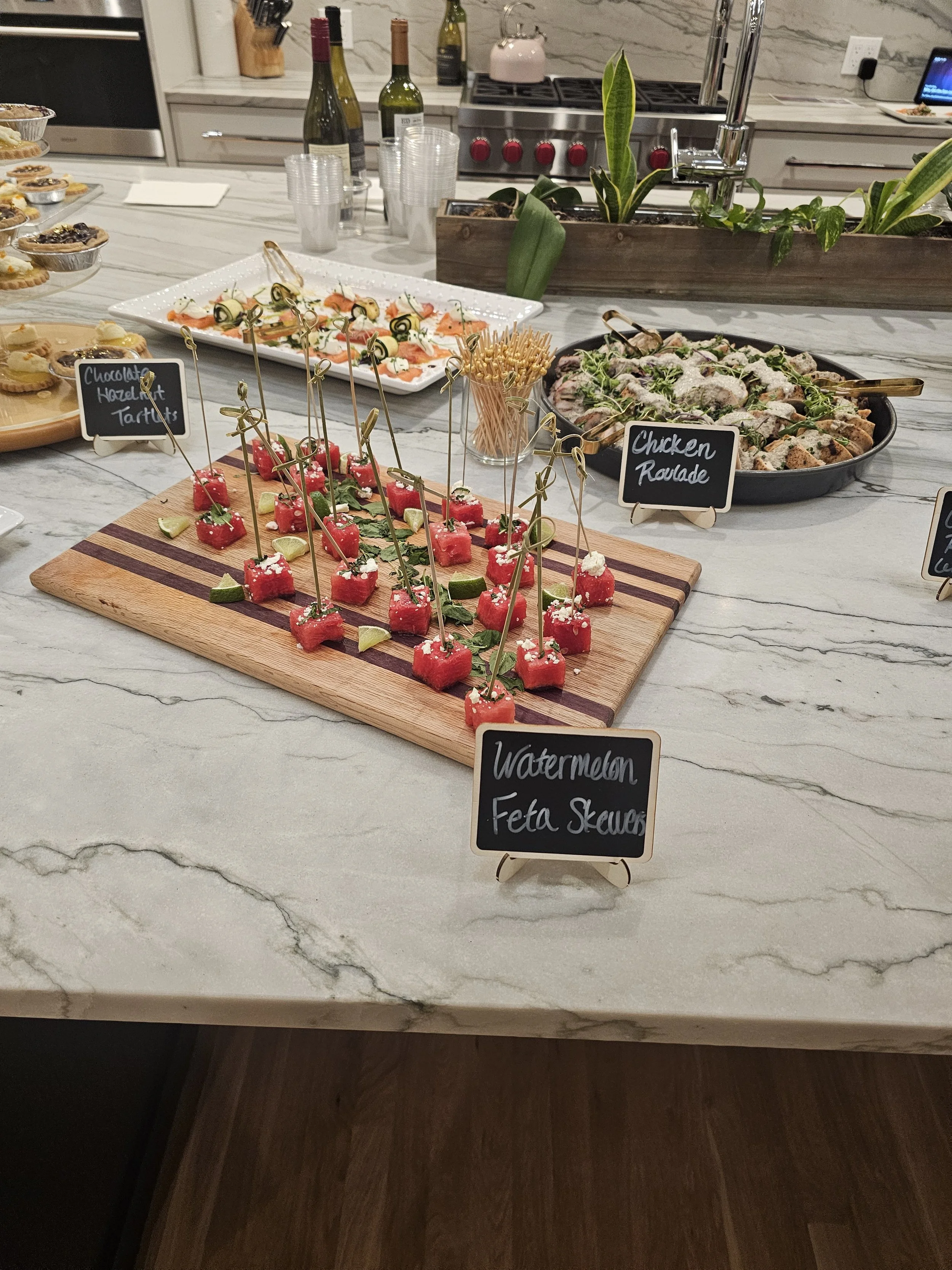 A buffet table with various appetizers including watermelon feta skewers, chicken roulade, and other finger foods, with chalkboard signs labeling the dishes, set on a marble countertop.