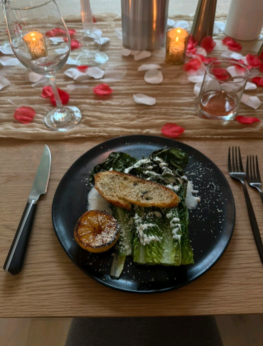 A plate of grilled romaine lettuce with a lemon half, a slice of toasted bread, and grated cheese on a dark plate. The table has a rustic wooden surface with a beige table runner, rose petal decorations, candles, and glassware nearby.