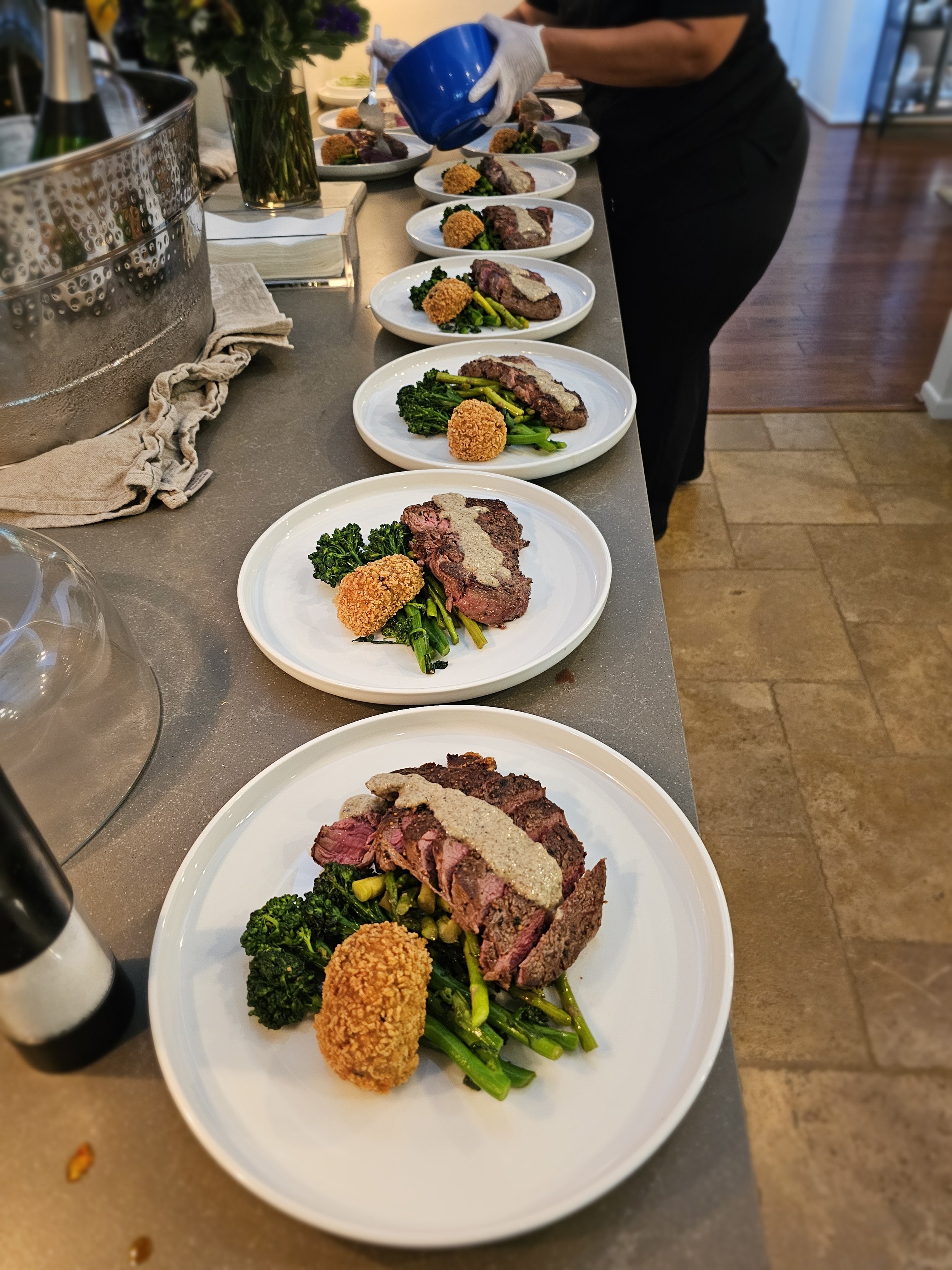 Several plates of cooked steak topped with sauce, served with green vegetables and a breaded side item on each plate, arranged on a counter.