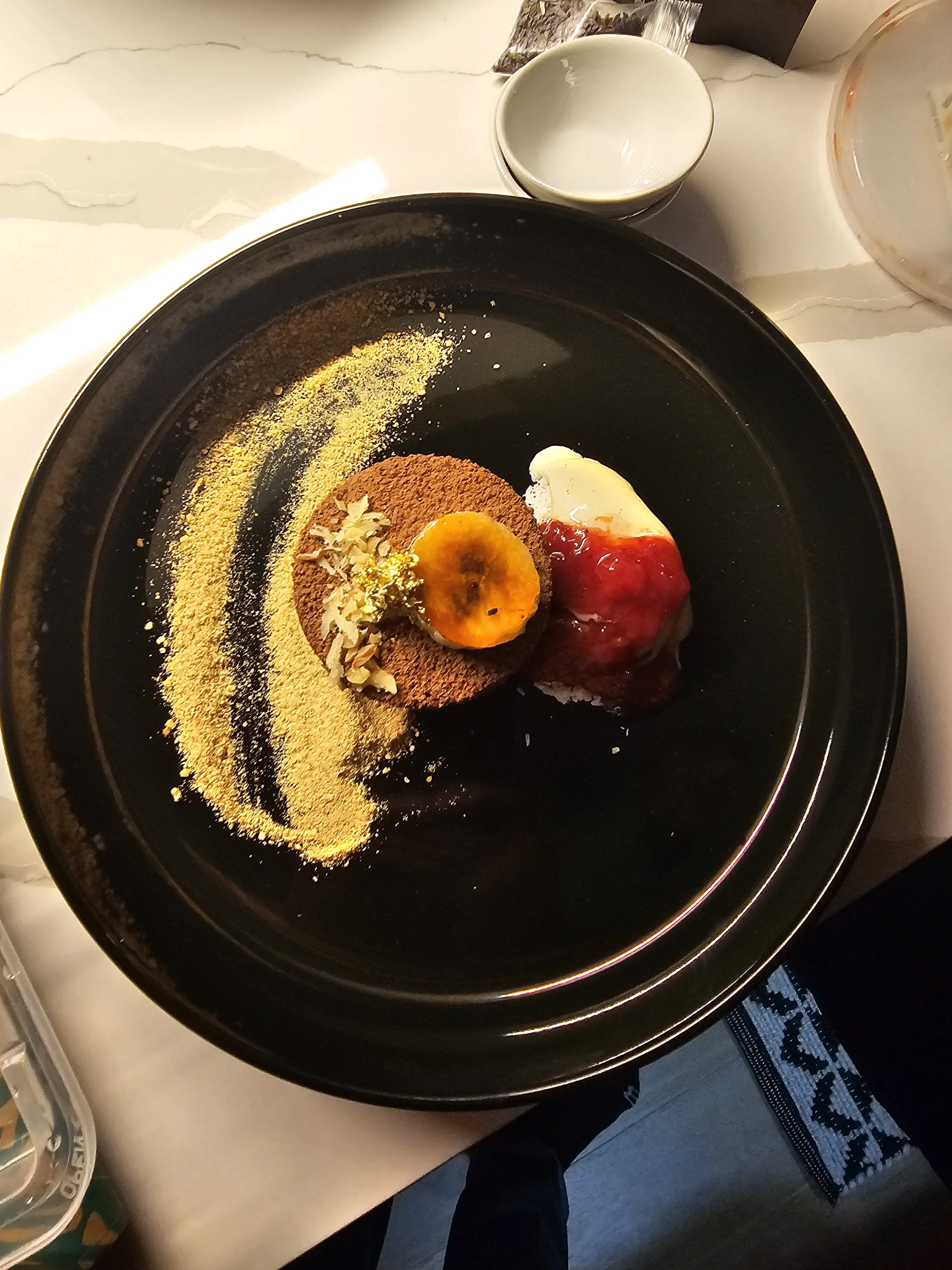 A black plate with a dessert that includes sliced banana, whipped cream, and red jelly, decorated with chopped nuts and yellow powder. There is also a faint yellow powder pattern on the plate. A white bowl and a teacup are visible on the white table 
