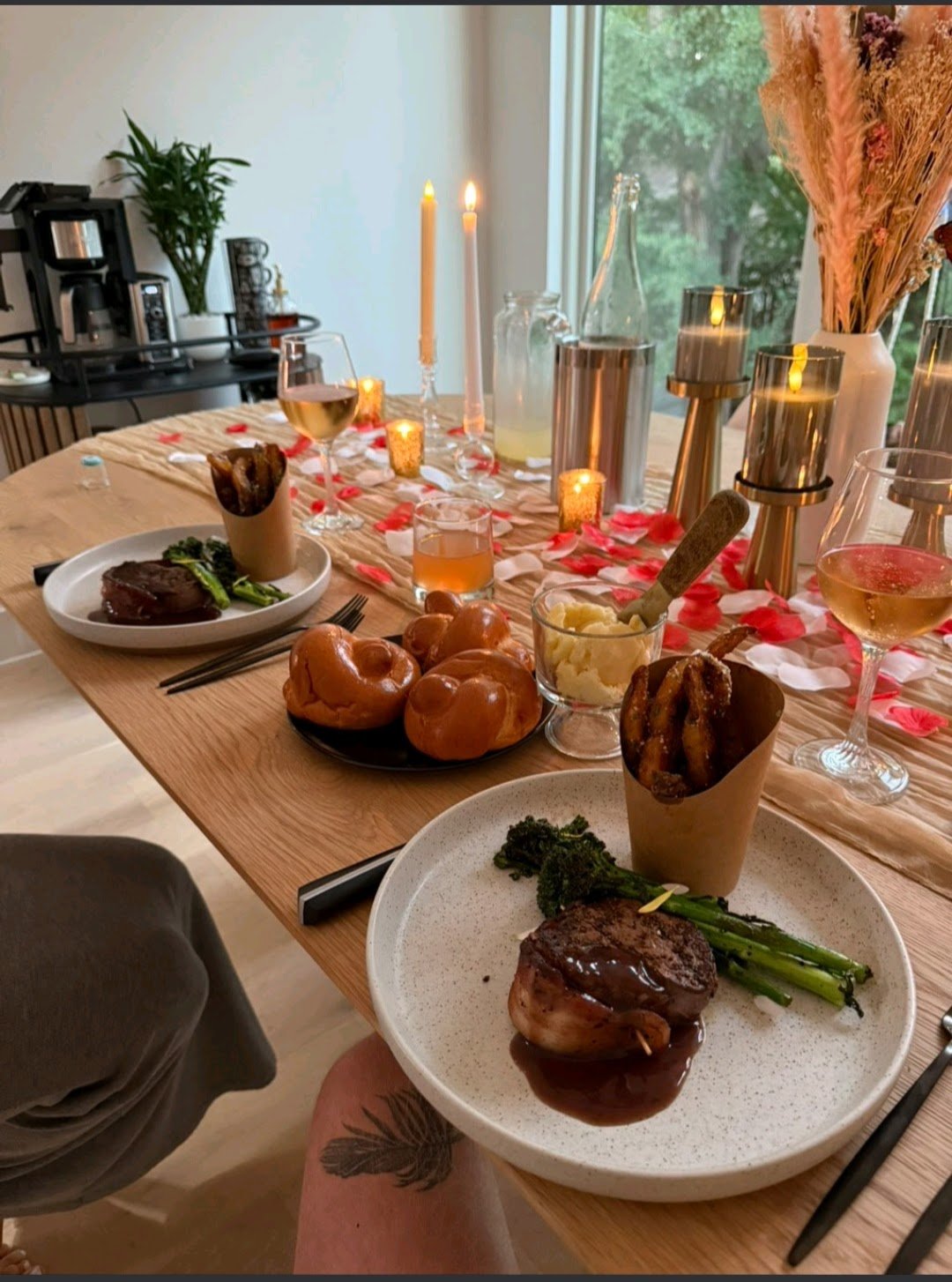 A private couples dinner set with candles, wine glasses, bread rolls, filet mignon, brocolini, mashed potatoes, and parmesan truffle fries, with a window and greenery outside.
