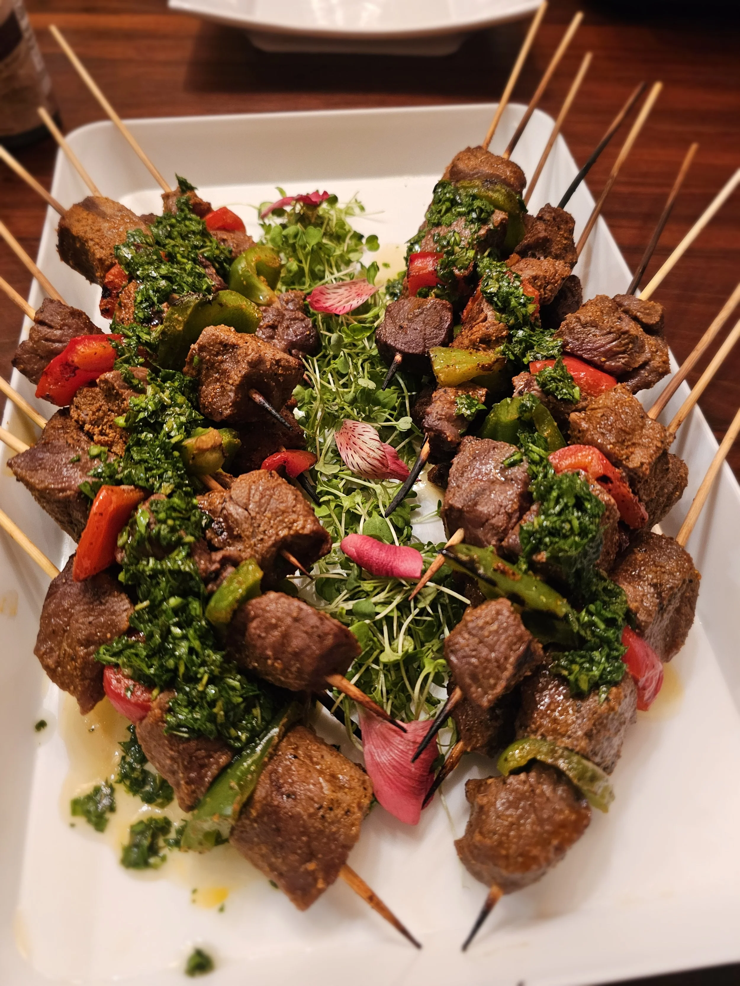 Skewers of grilled beef with green peppers and chopped herbs, garnished with microgreens and edible flower petals on a white rectangular plate.