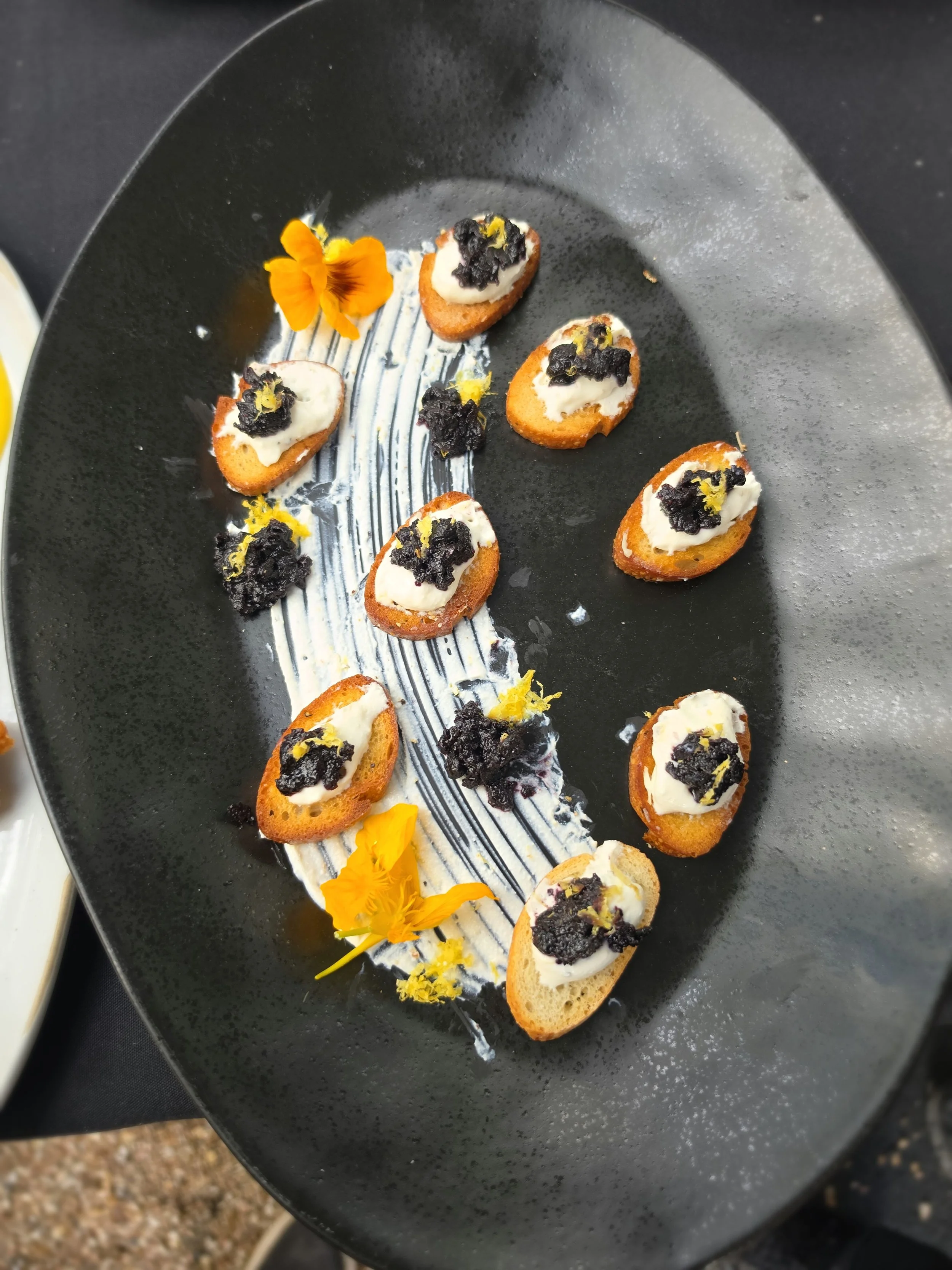Small toast slices topped with a dollop of creamy spread, black crumbs, and a yellow garnish, garnished with bright orange edible flowers, arranged on a black plate with a decorative stripe.