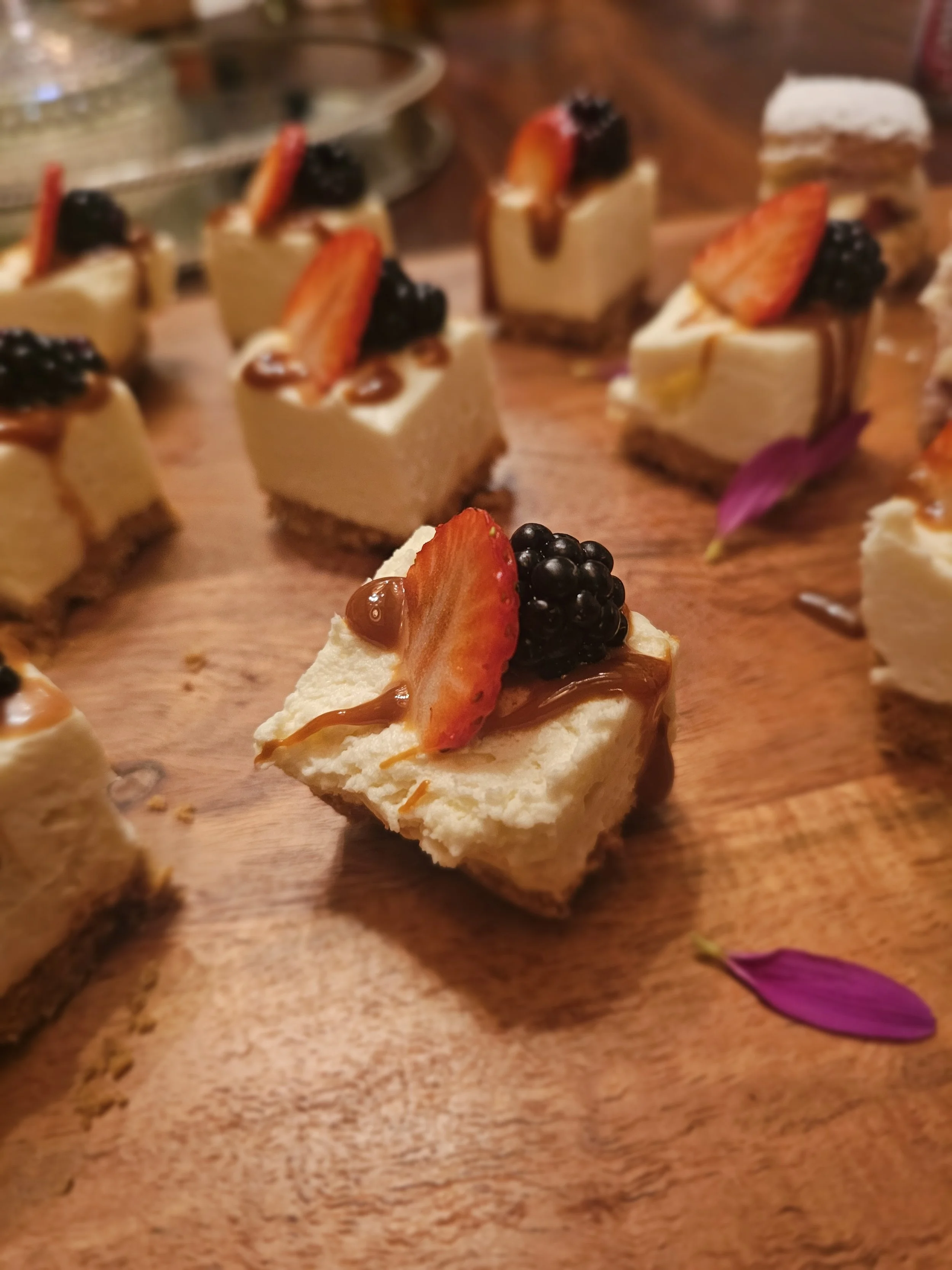 Small dessert topped with sliced strawberries, blackberries, caramel, and a purple flower on a wooden surface.