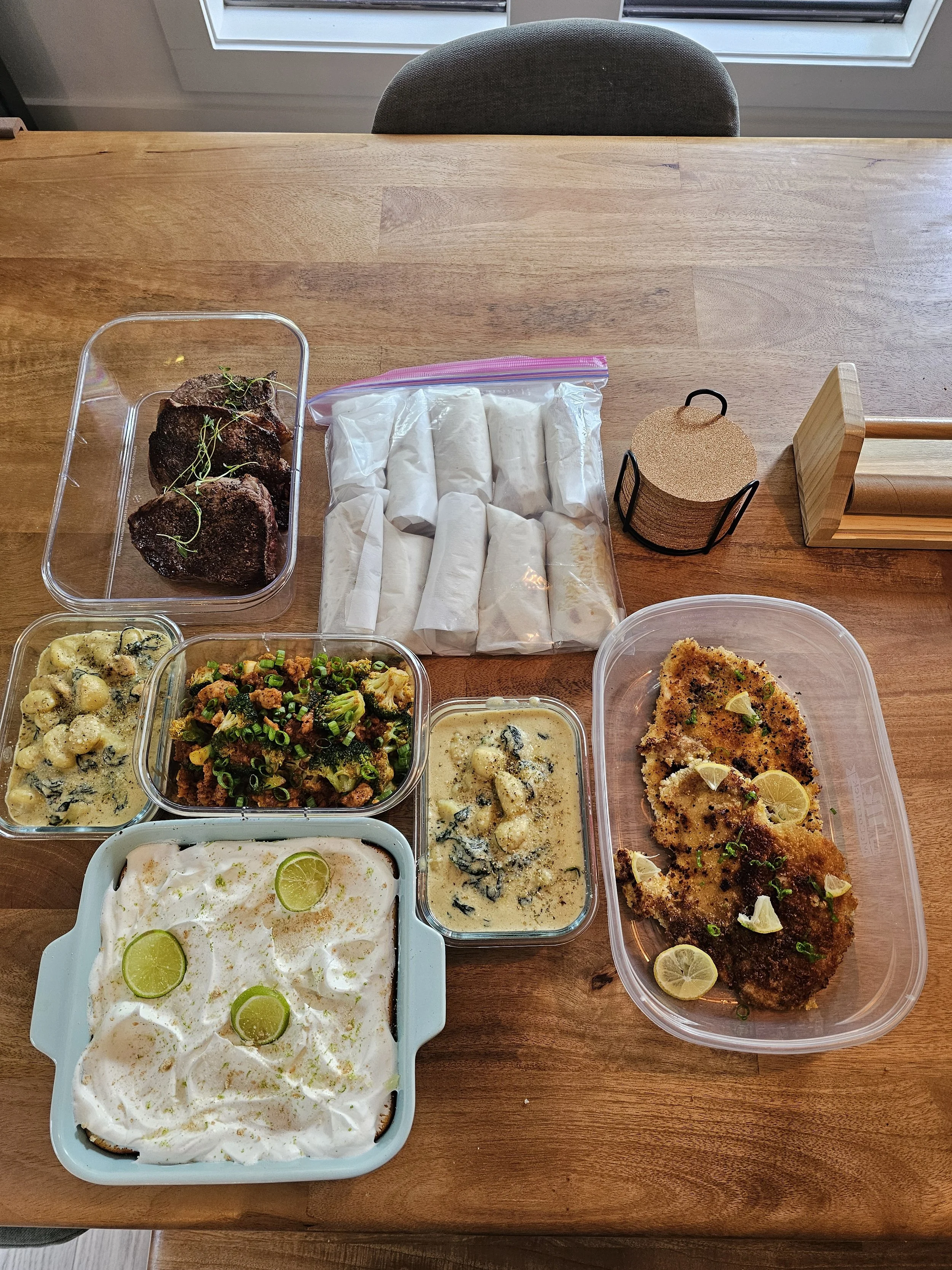 A wooden table with various dishes of food including baked chicken, rolls, a salad, potato salad, a casserole with broccoli, a creamy dish, and fried fish, with a cup of coasters and a baking dish, in a well-lit room near a window.