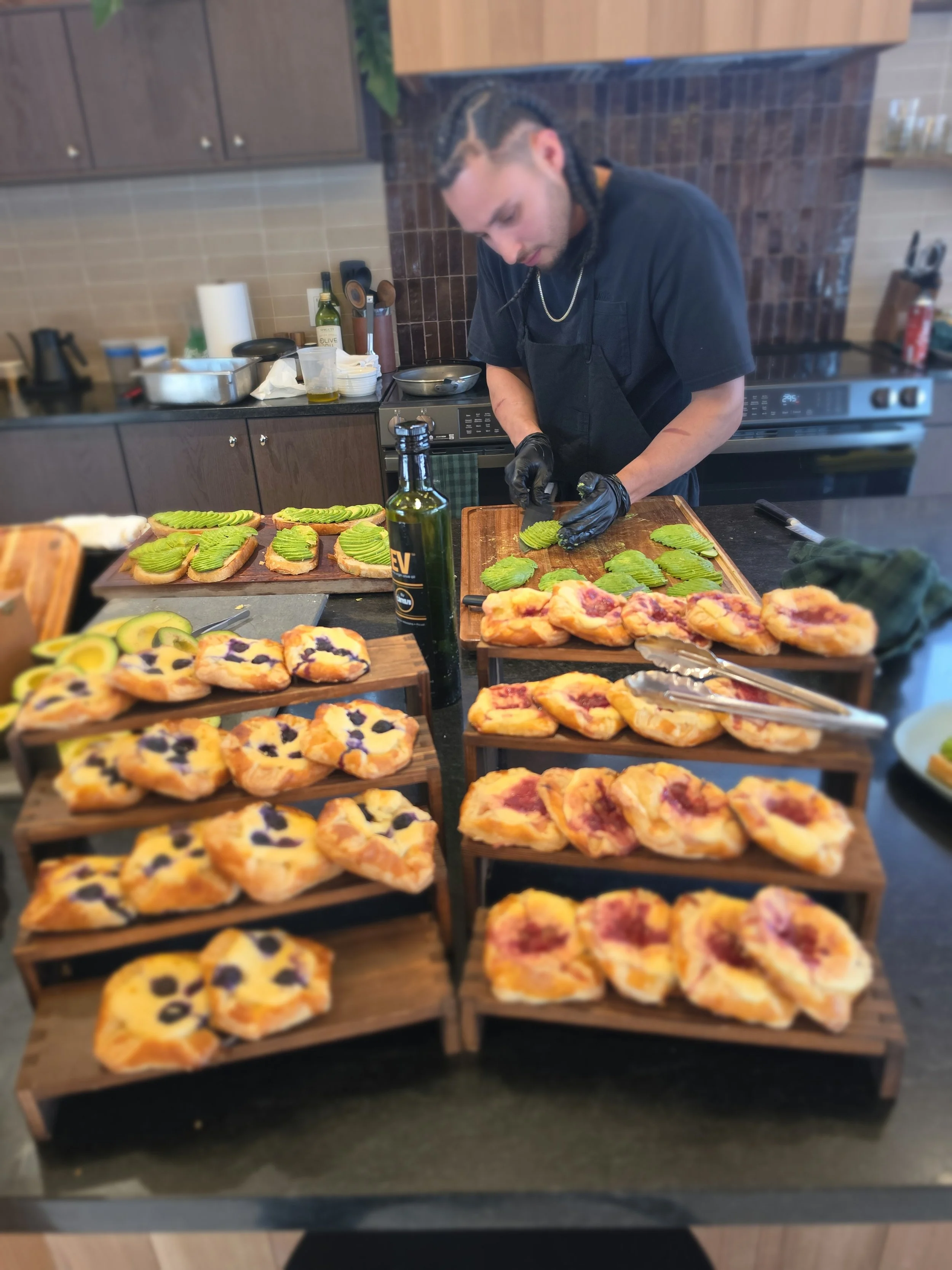 A man with braided hair wearing a black shirt and gloves preparing avocado toast and pizza slices on a wooden display stand in a modern kitchen with brown cabinets, a stove, and kitchen utensils in the background.