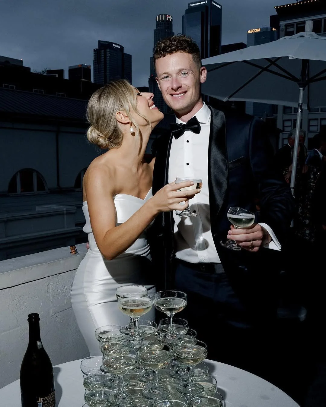 A couple dressed in formal attire at a rooftop party, with the woman in a white gown and the man in a tuxedo, holding wine glasses, surrounded by champagne glasses on the table, with city skyscrapers in the background.