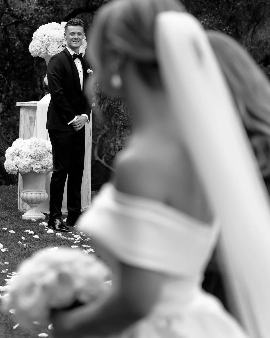 A groom in a tuxedo stands in front of an arch decorated with white flowers during a wedding ceremony. The image is in black and white, with a blurred bride in the foreground.