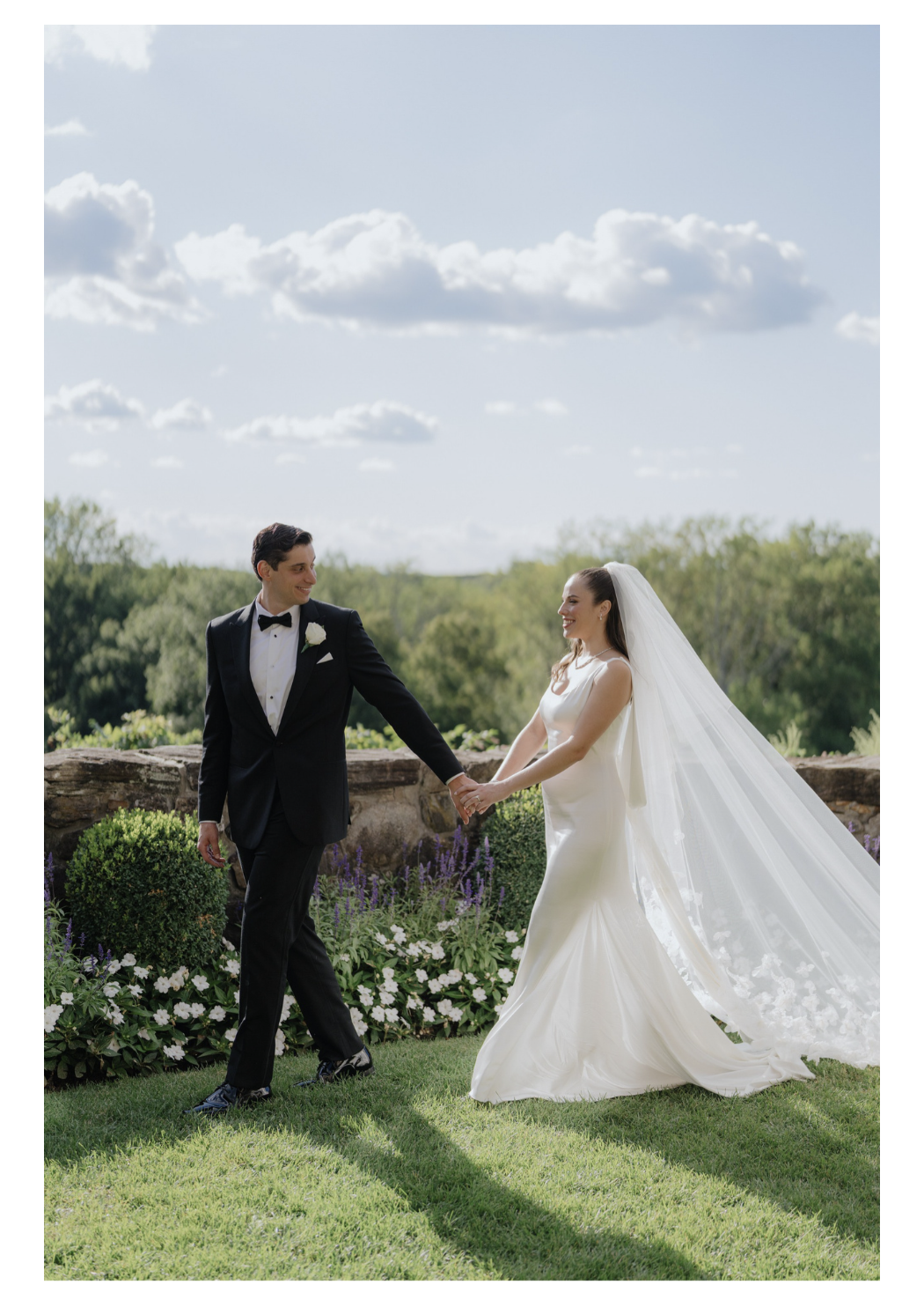 A bride and groom holding hands outdoors, smiling at each other, with a garden and blue sky with clouds in the background.