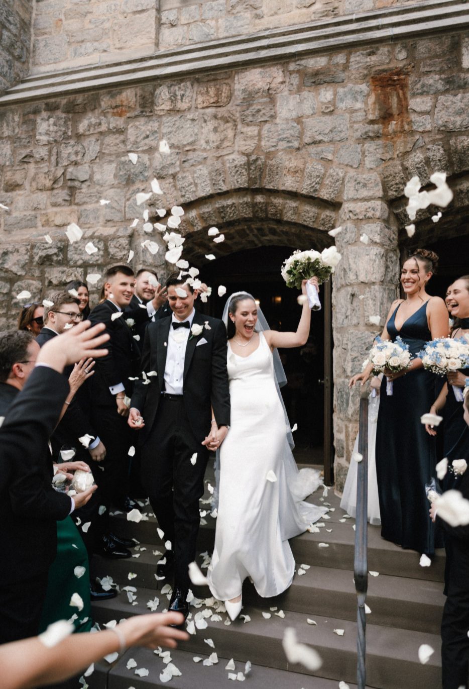 Bride and groom walking down stairs while guests throw flower petals at a wedding celebration outside a stone building.