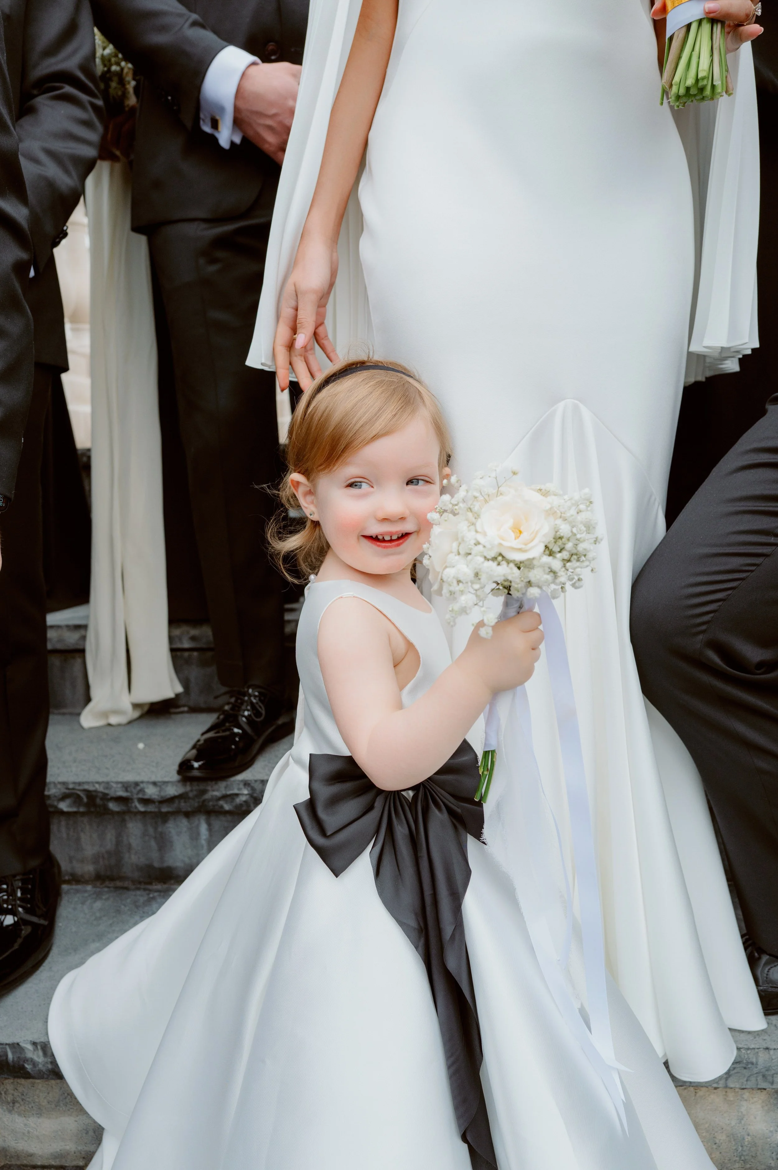 A young girl in a white dress with a black sash holding a bouquet of white flowers, standing on steps with adults dressed in formal black attire nearby.