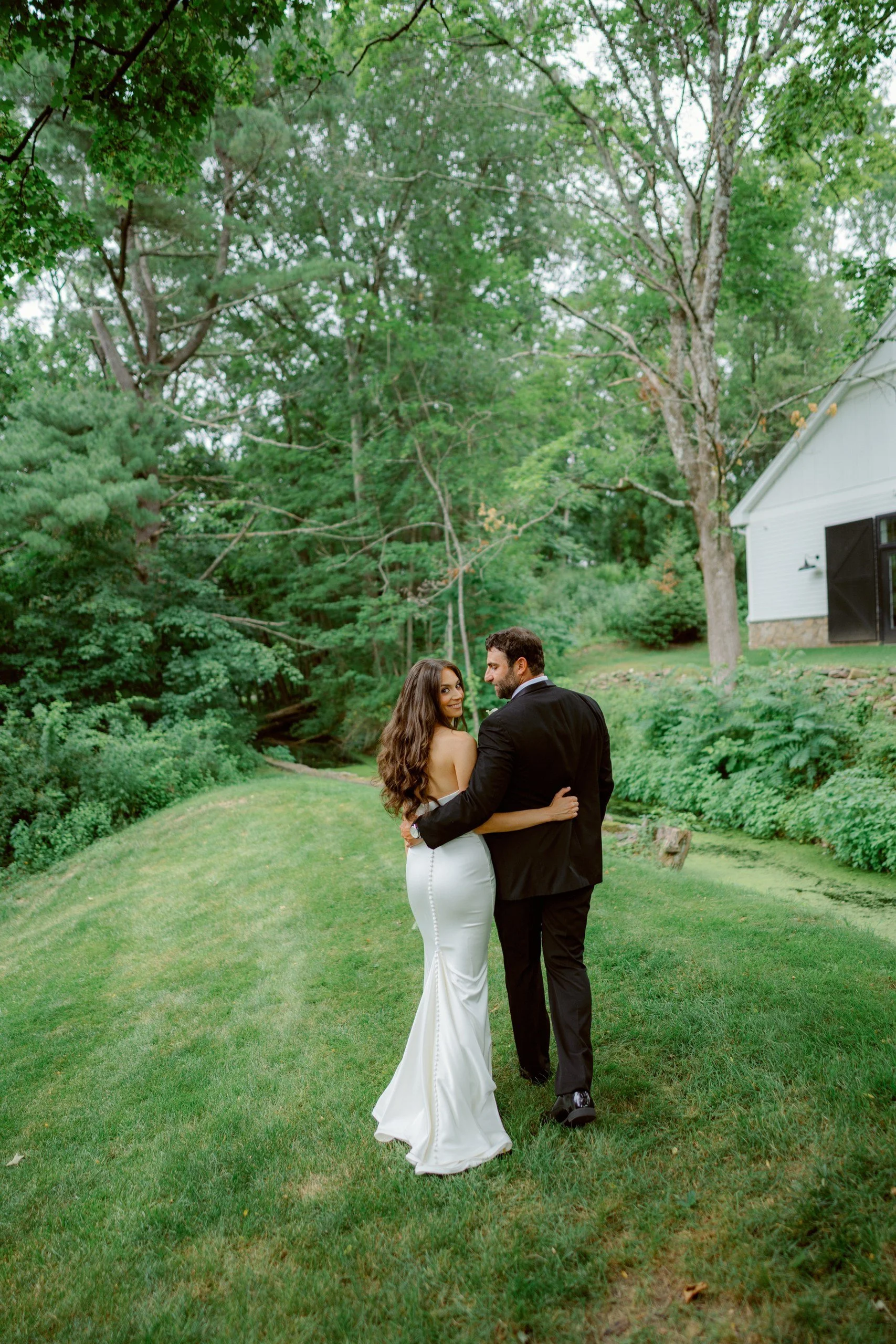 A newlywed couple standing on a grassy lawn, embracing each other under a large tree with a white house visible in the background, surrounded by greenery.
