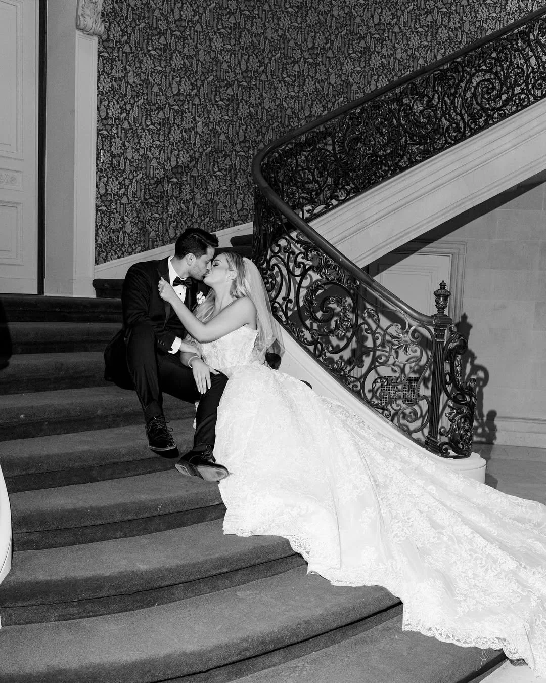 A couple in wedding attire sharing a kiss on a staircase with ornate railing, black and white photo.