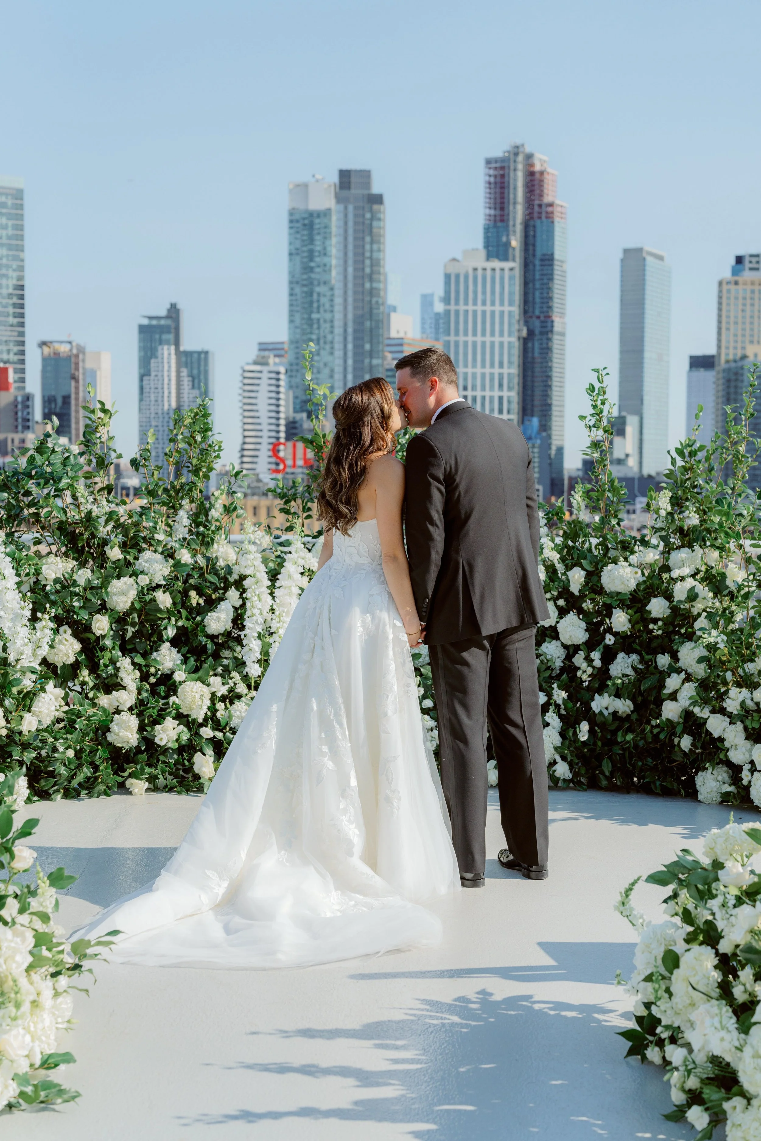 A bride and groom kissing on a rooftop with a city skyline in the background, surrounded by white flowers and greenery.