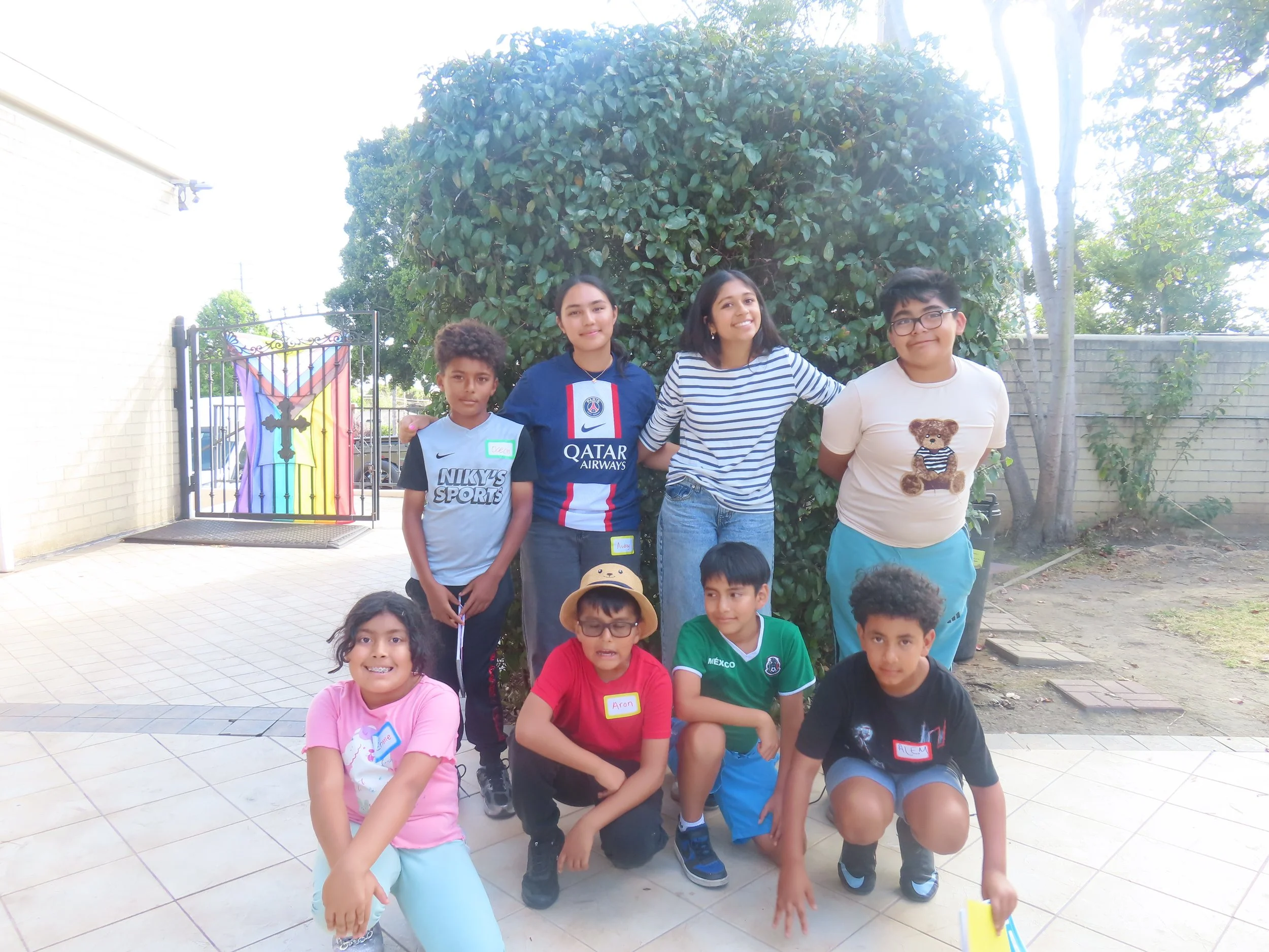 A group of children and teenagers outdoors near a large bush and a brick wall, some wearing name tags. They are smiling and posing for a photo.